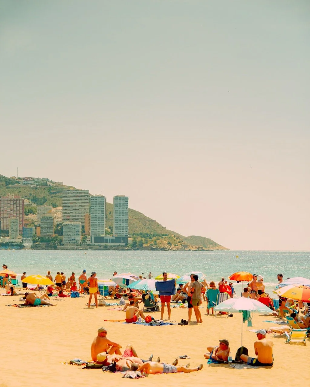 Playa con muchas personas, sombrillas y arena, con edificios y colinas en el fondo.