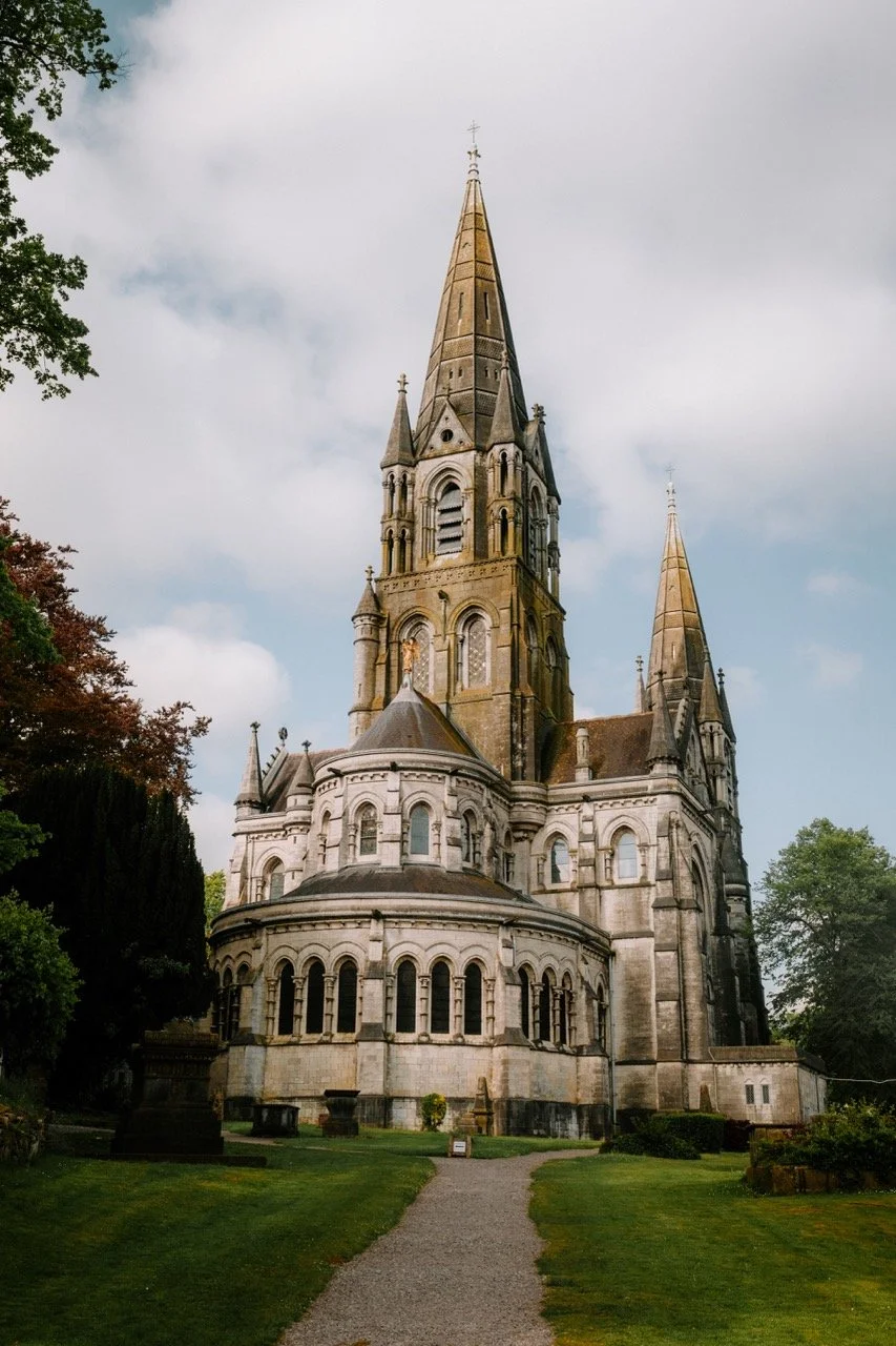 Catedral gótica con torres altas, ventanas arqueadas y un camino de piedra que lleva hacia ella, rodeada de césped y árboles.