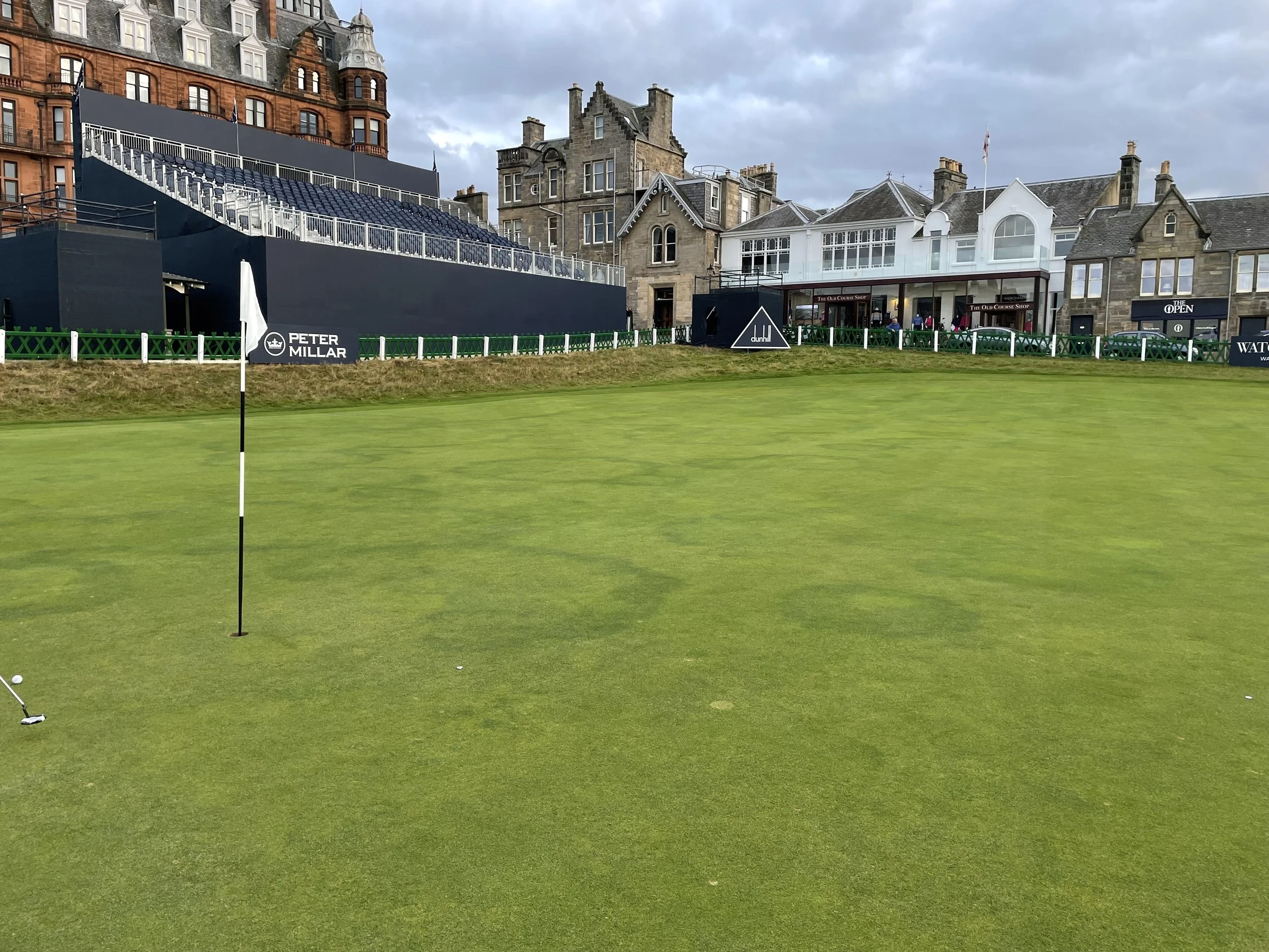 A golf course with a putting green, flagstick, and two golf balls in the foreground. In the background, there are historic buildings, a viewing stand or bleachers, and people near a clubhouse with signage for Peter Millar and other brands.