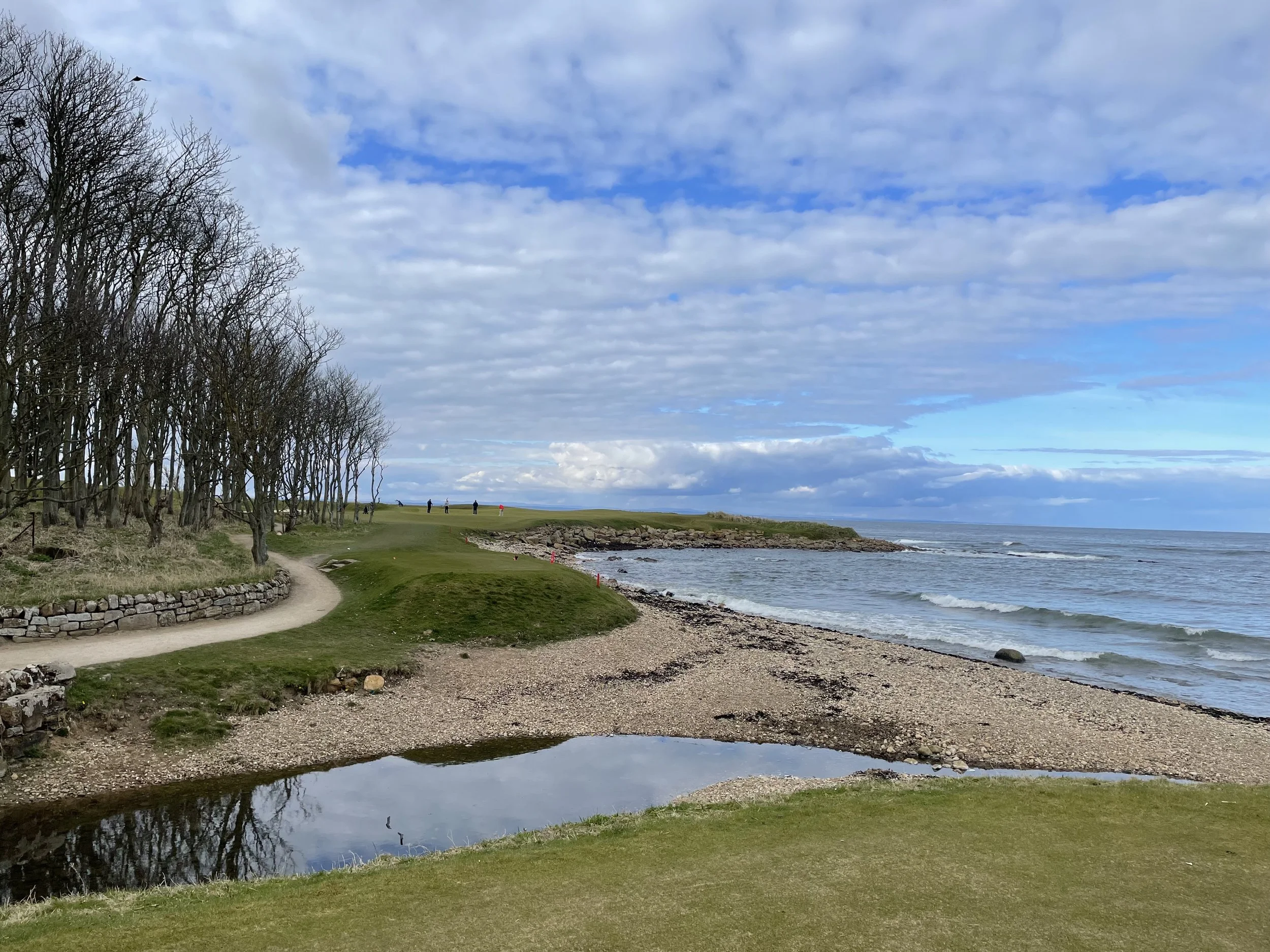 Coastal scene of a golf course with a walking path, trees on the left, a pebble beach, calm ocean waves on the right, and a partly cloudy sky above.