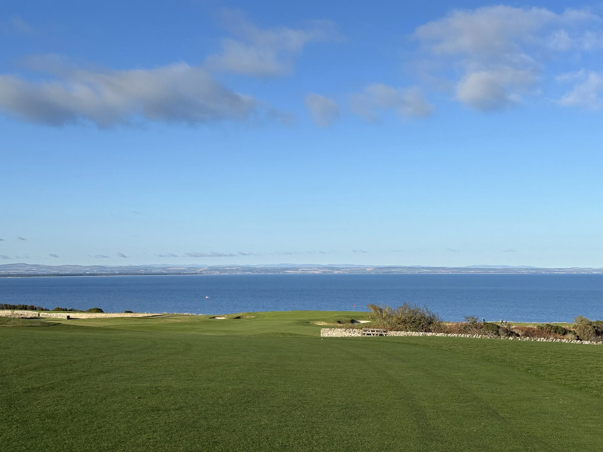 Golf course with green grass, coastline view, ocean, and partly cloudy sky.