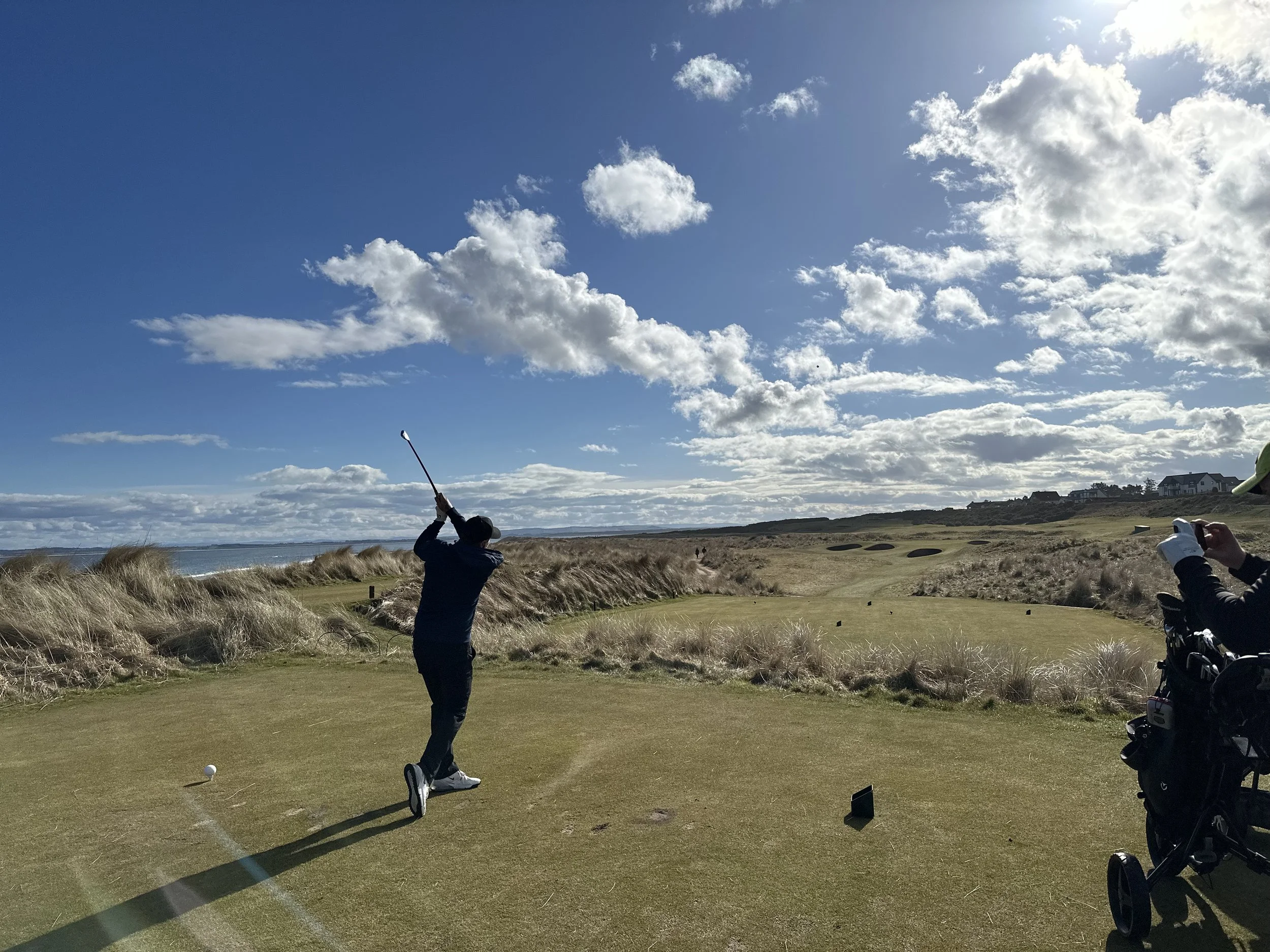 A person playing golf on a grassy course with sand dunes and a partly cloudy sky in the background.