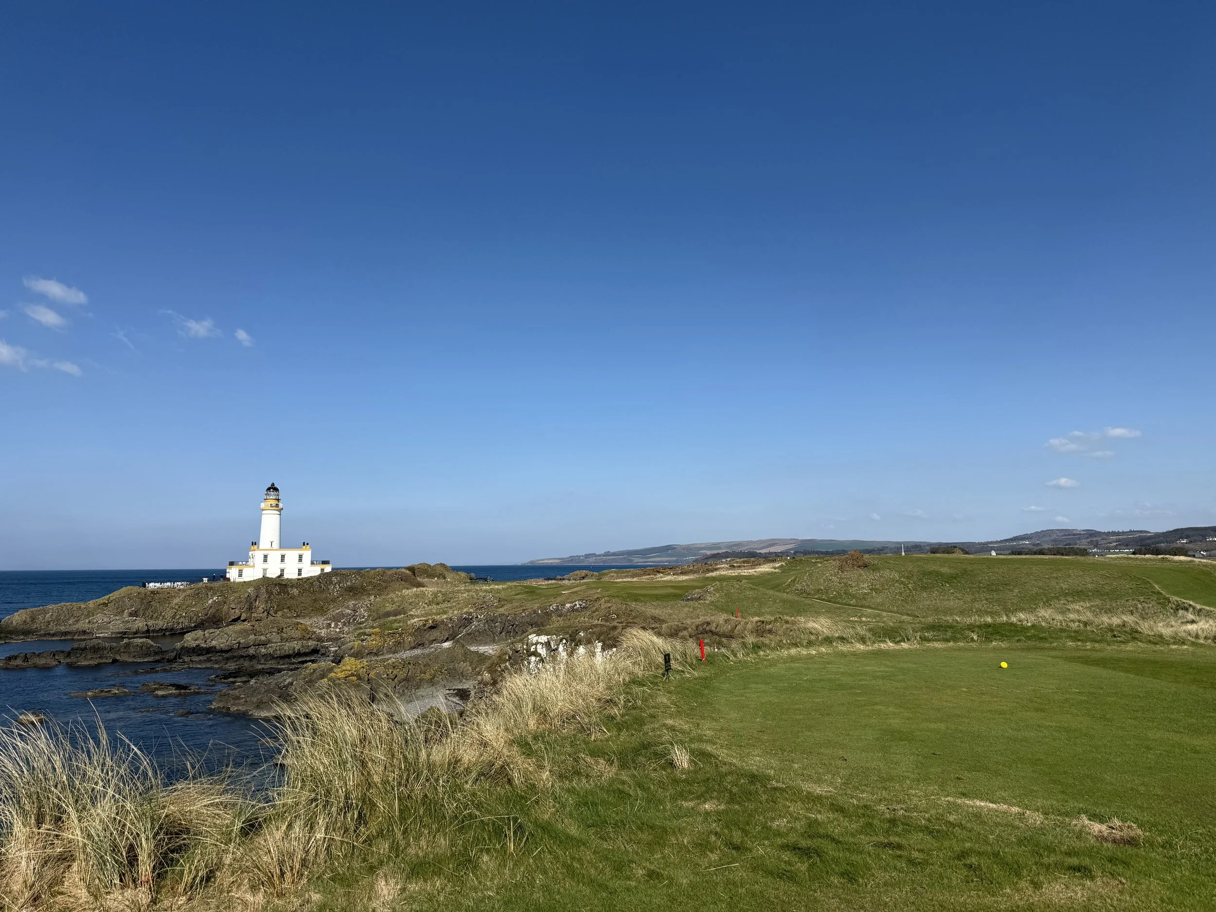 A lighthouse on a rocky coastline with a golf course in the foreground and a clear blue sky.