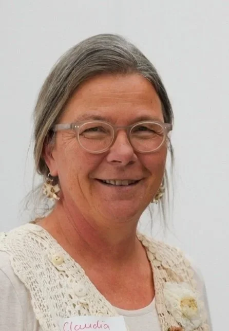A woman with gray hair, glasses, and earrings, smiling in front of a plain white background.