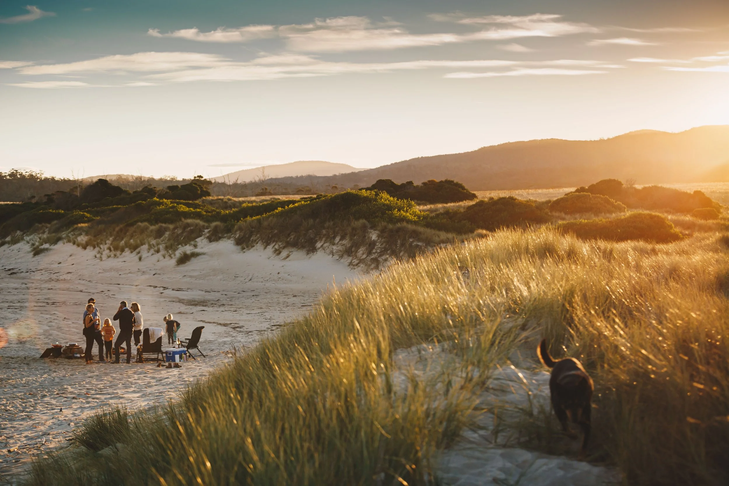 Family gathering on the beach at sunset, with chairs and food set up, tall grass in the foreground, and mountains in the background.