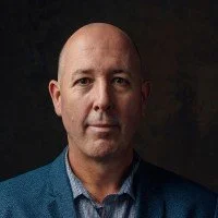 A portrait of a bald man in a blue blazer against a dark background.