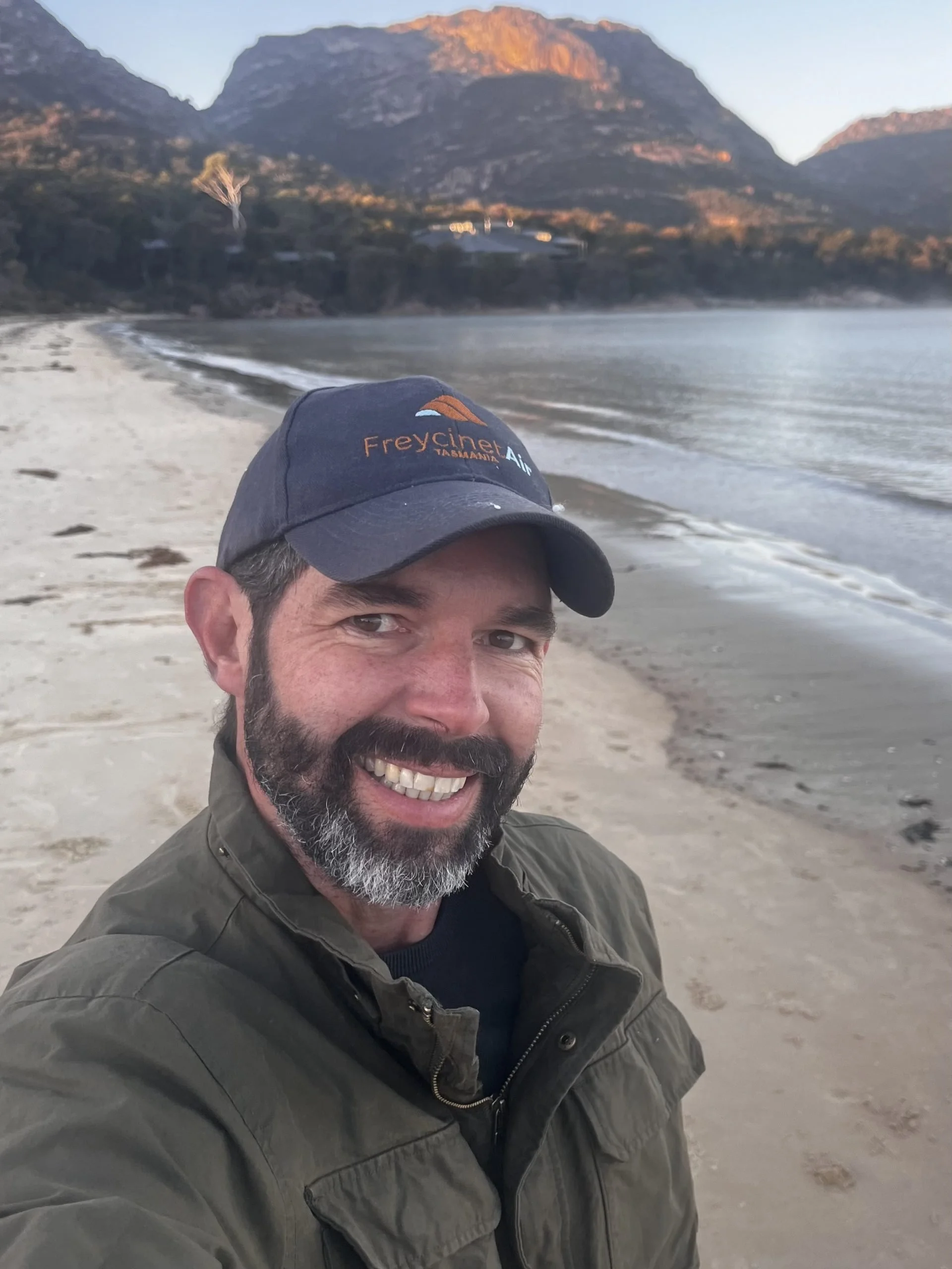 A man smiling with gray beard and hair taking a selfie at the beach during sunset, wearing a blue cap and green jacket. Mountains and water are visible in the background.