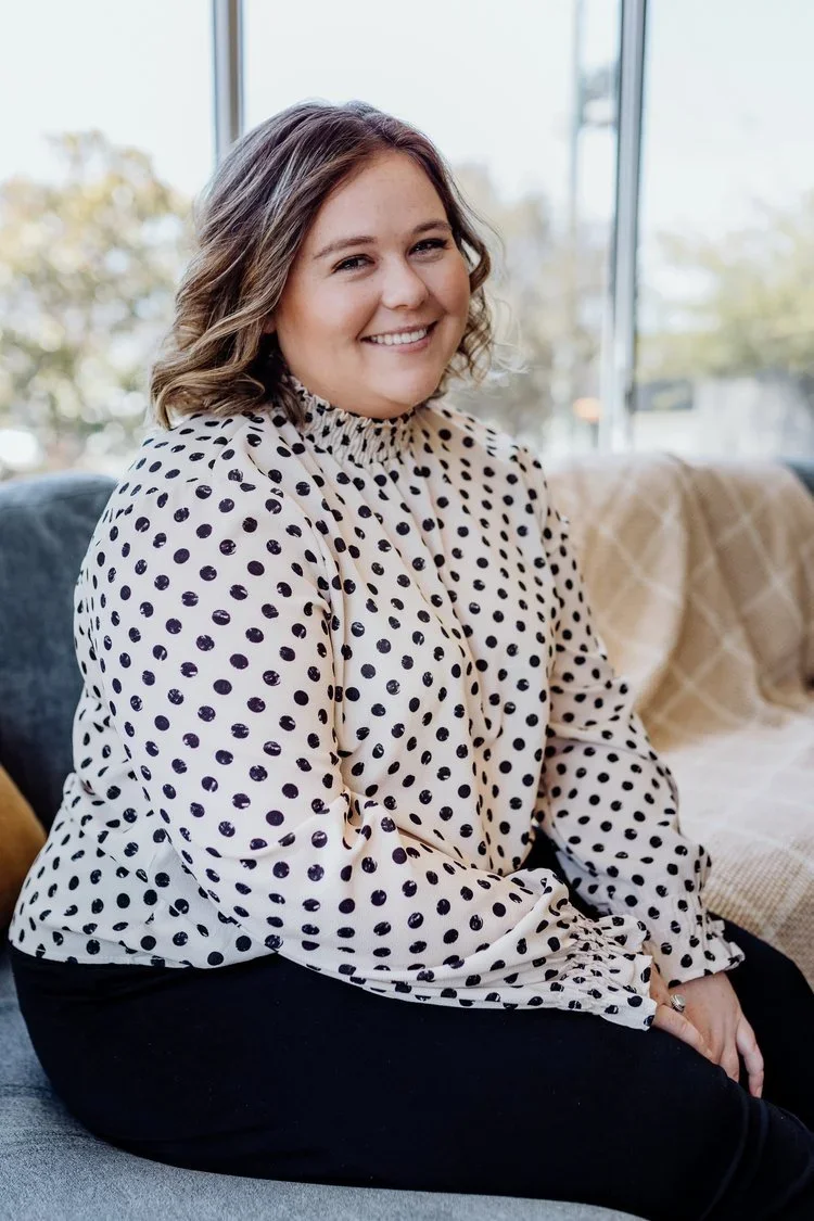 A woman with shoulder-length curly hair sitting on a sofa, smiling, wearing a white blouse with black polka dots and black pants, with a window and outdoor trees in the background.