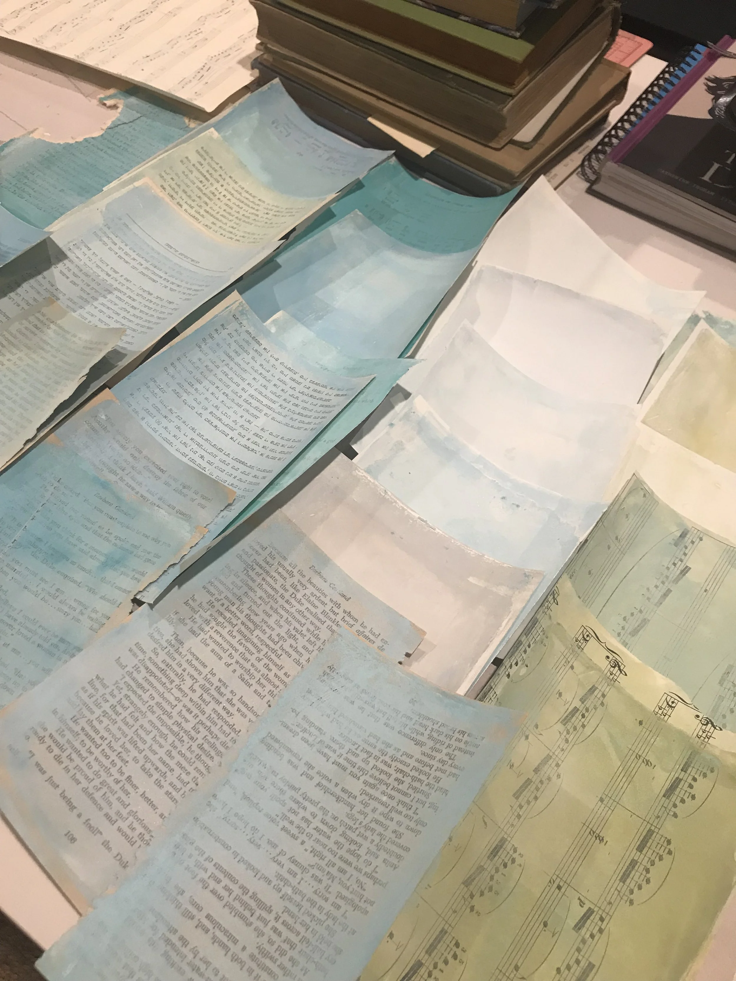 A collection of custom colored collage papers made from book pages, found papers, and musical scores scattered across a table.