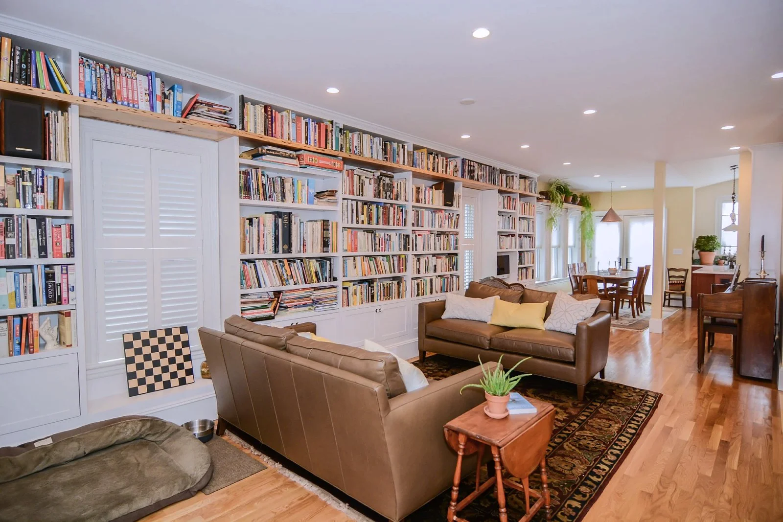 Living room with a large white bookshelf wall, brown leather sofas, a wooden side table with a potted plant, and a dining area in the background.
