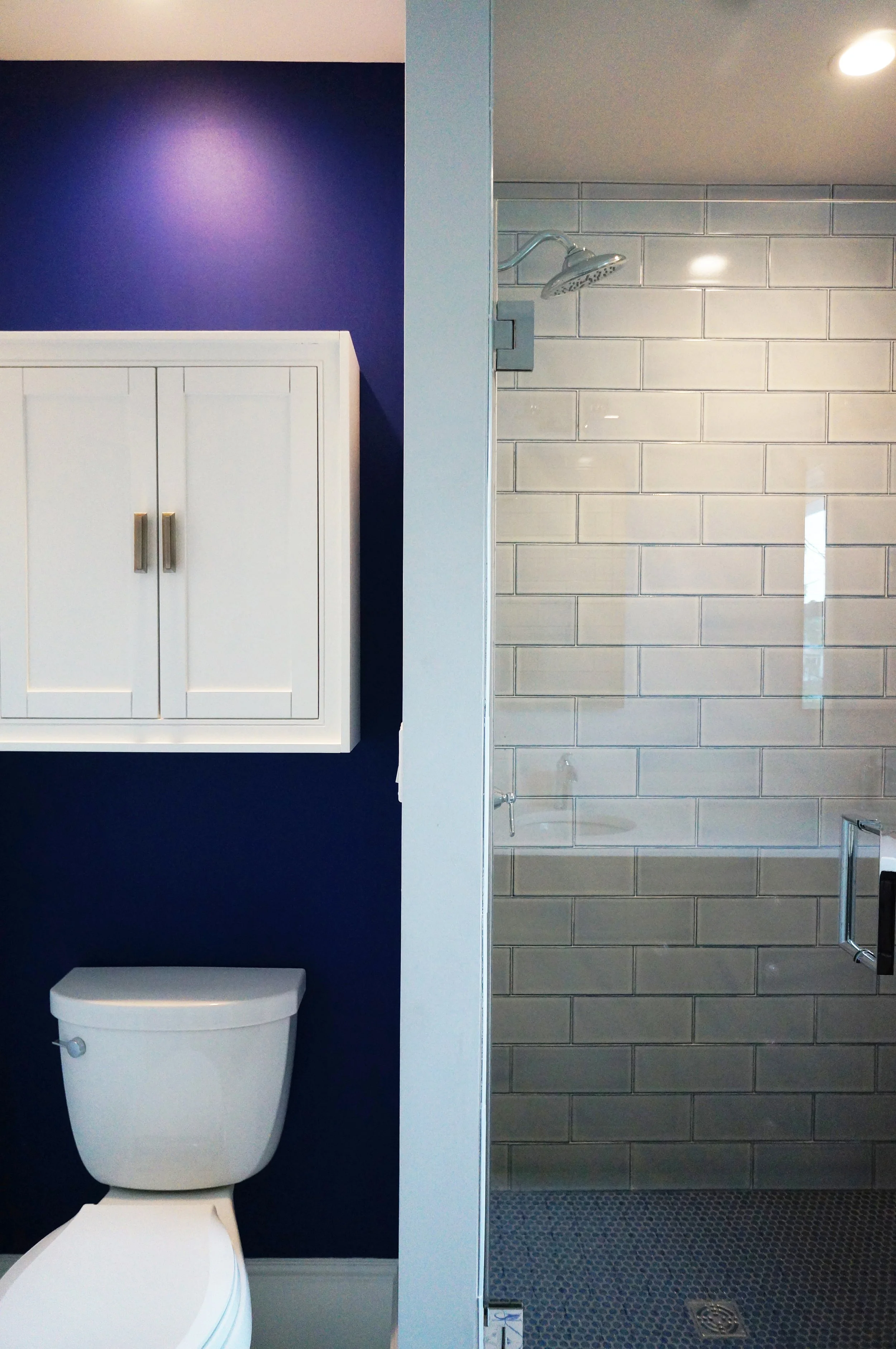 A bathroom with a blue wall and a white toilet on the left, and a walk-in shower with a glass door and gray subway tiles on the right.