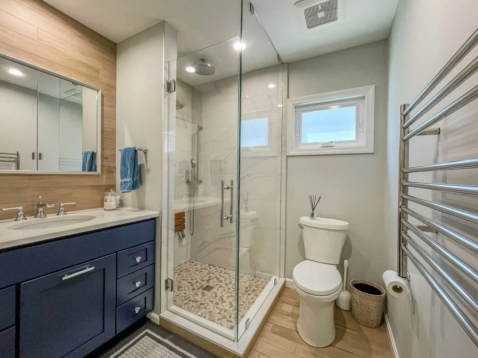 Modern bathroom with a glass shower enclosure, blue vanity with double sinks, mirror, towel racks, a toilet, small window, and wooden floor.