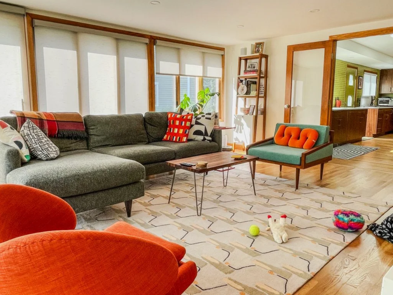 Living room with a gray sectional sofa with colorful throw pillows, a wooden coffee table, and a teal armchair with an orange pillow. Large windows with blinds, a patterned area rug, and a bookshelf filled with books and pictures. The room opens into a kitchen with wooden cabinets and a striped rug on the hardwood floor. Toys including a tennis ball, plush rabbit, and a colorful toy bowl are on the rug.