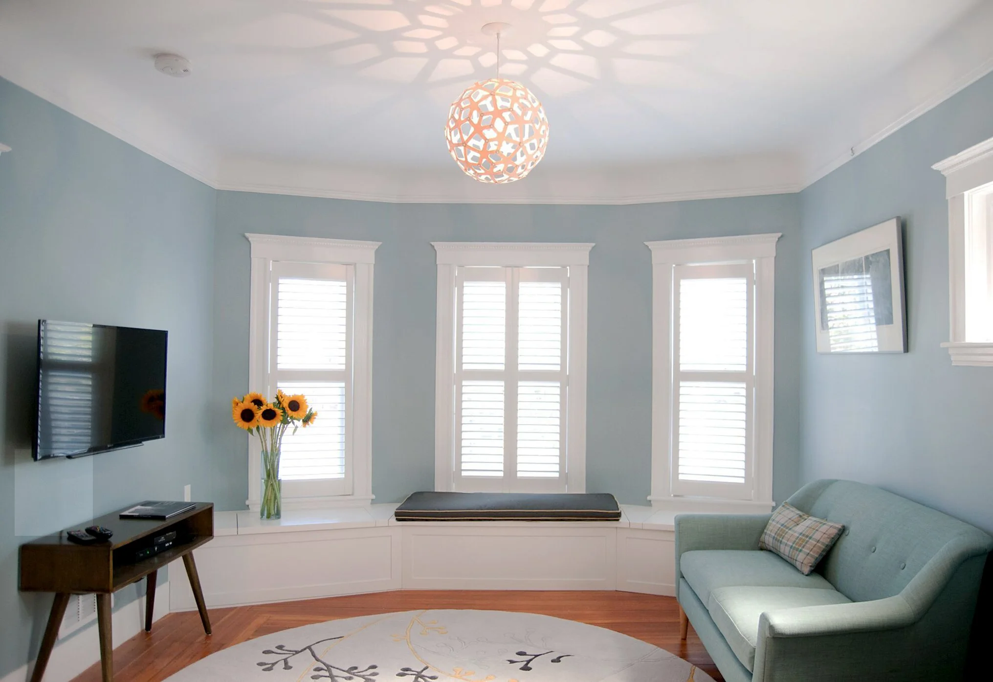 Living room with light blue walls, white window shutters, a modern ceiling light casting a shadow pattern, a flat-screen TV on the wall, a small wooden table with a remote control, a vase of sunflowers, a window seat with a cushion, a light green sof