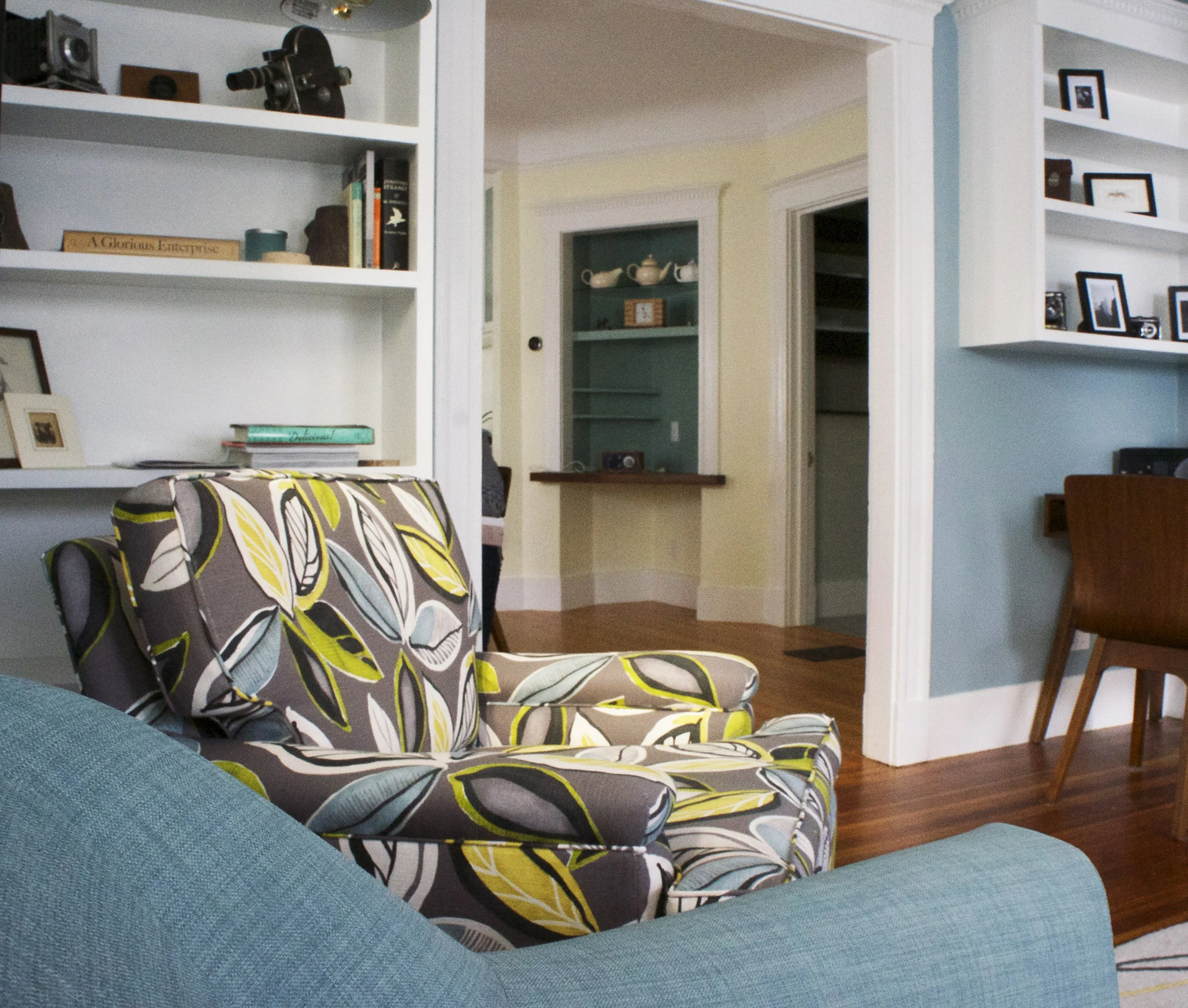 Living room with patterned armchair, blue sofa, white bookshelves with framed photos and books, wooden floor, doorway leading to other rooms.