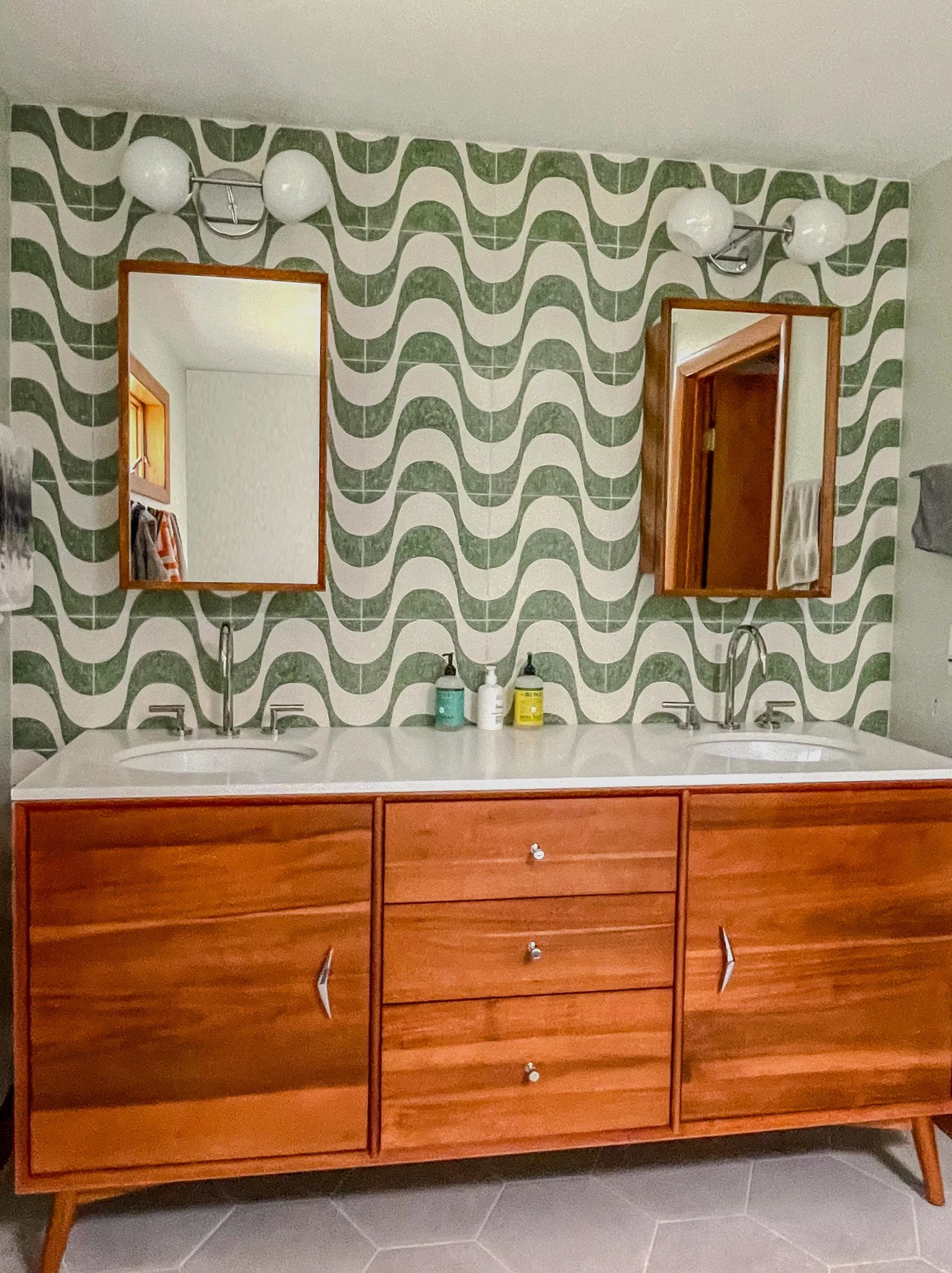 A bathroom vanity with twin sinks, two mirrors, and a patterned green and white wallpaper.