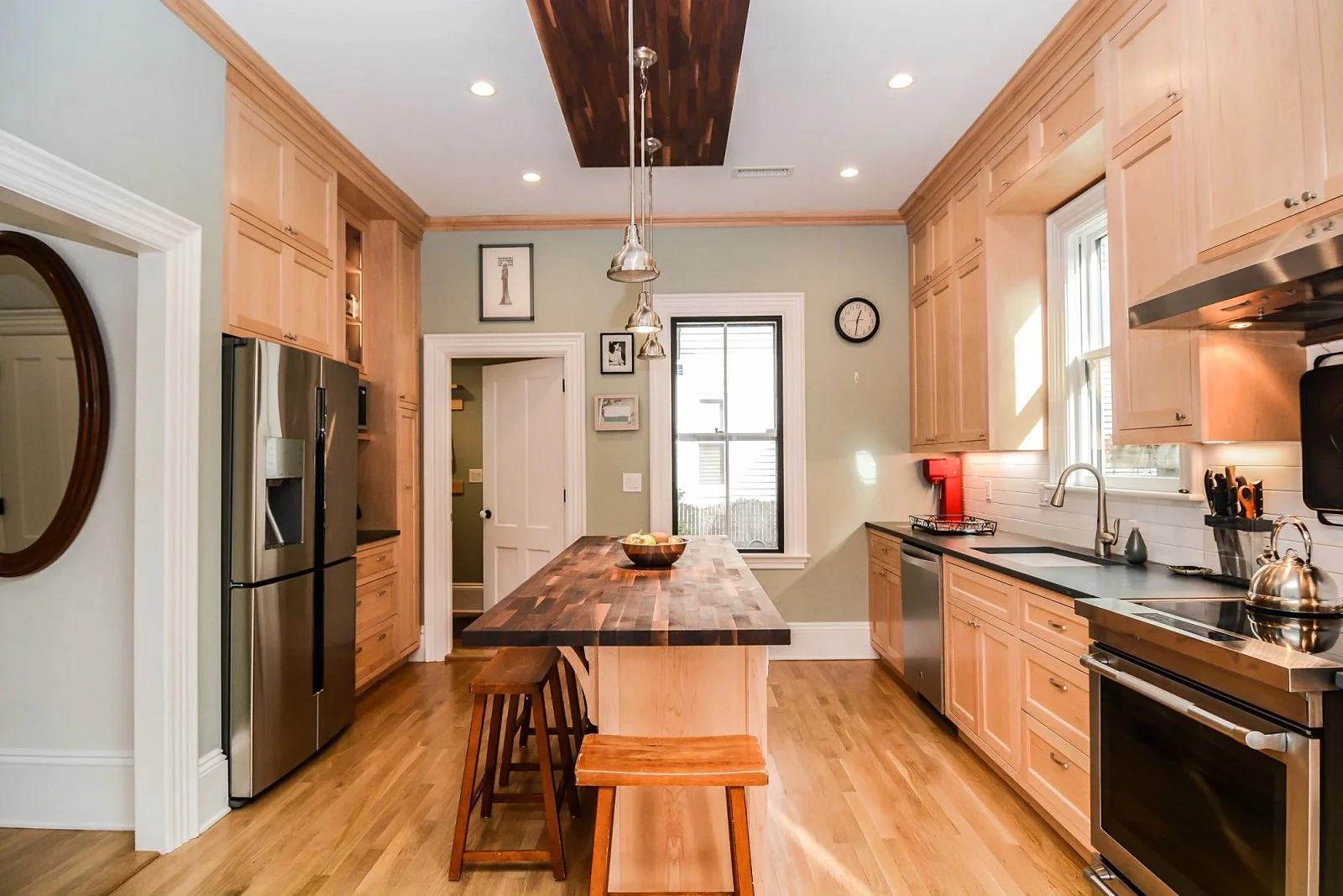 A modern kitchen with light wood cabinetry, stainless steel appliances including a refrigerator and oven, a large wooden center island with stools, hardwood floors, and a window allowing sunlight in.