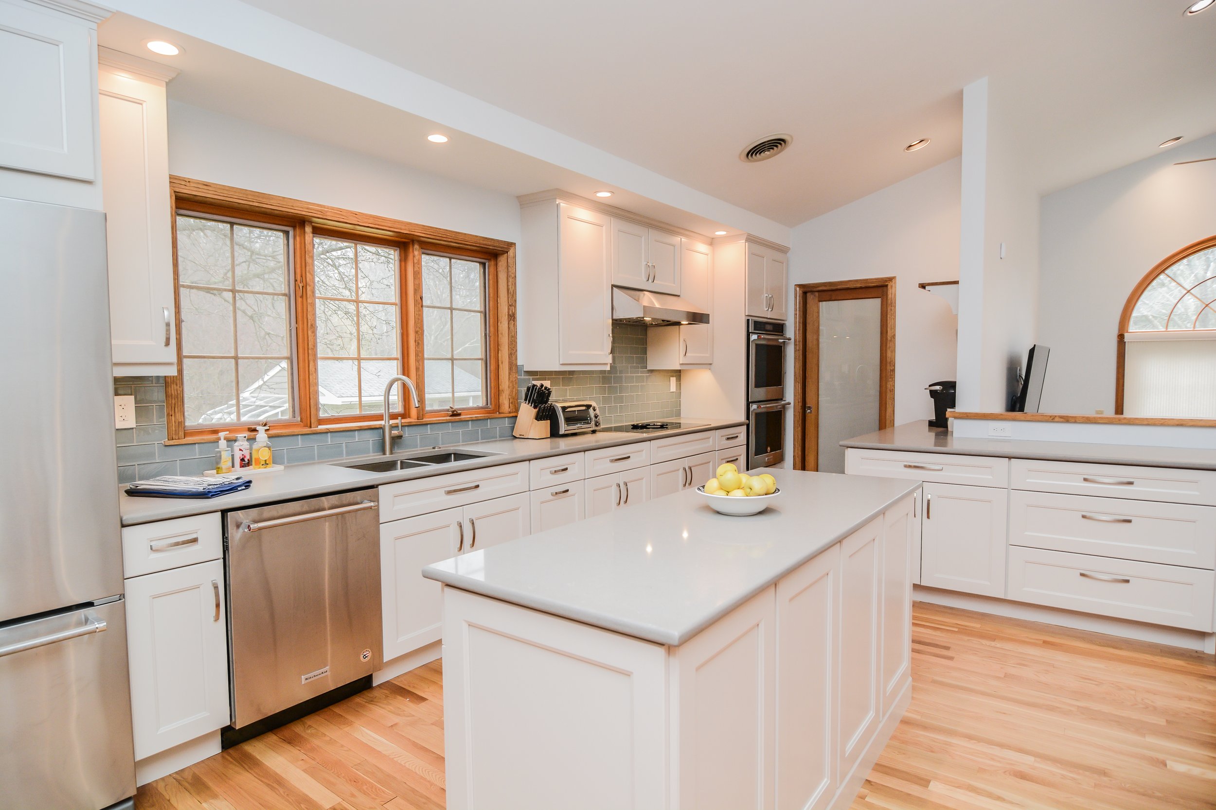 Bright kitchen with white cabinets, a kitchen island with a bowl of yellow apples, large window with wooden trim, hardwood floors, and modern appliances.