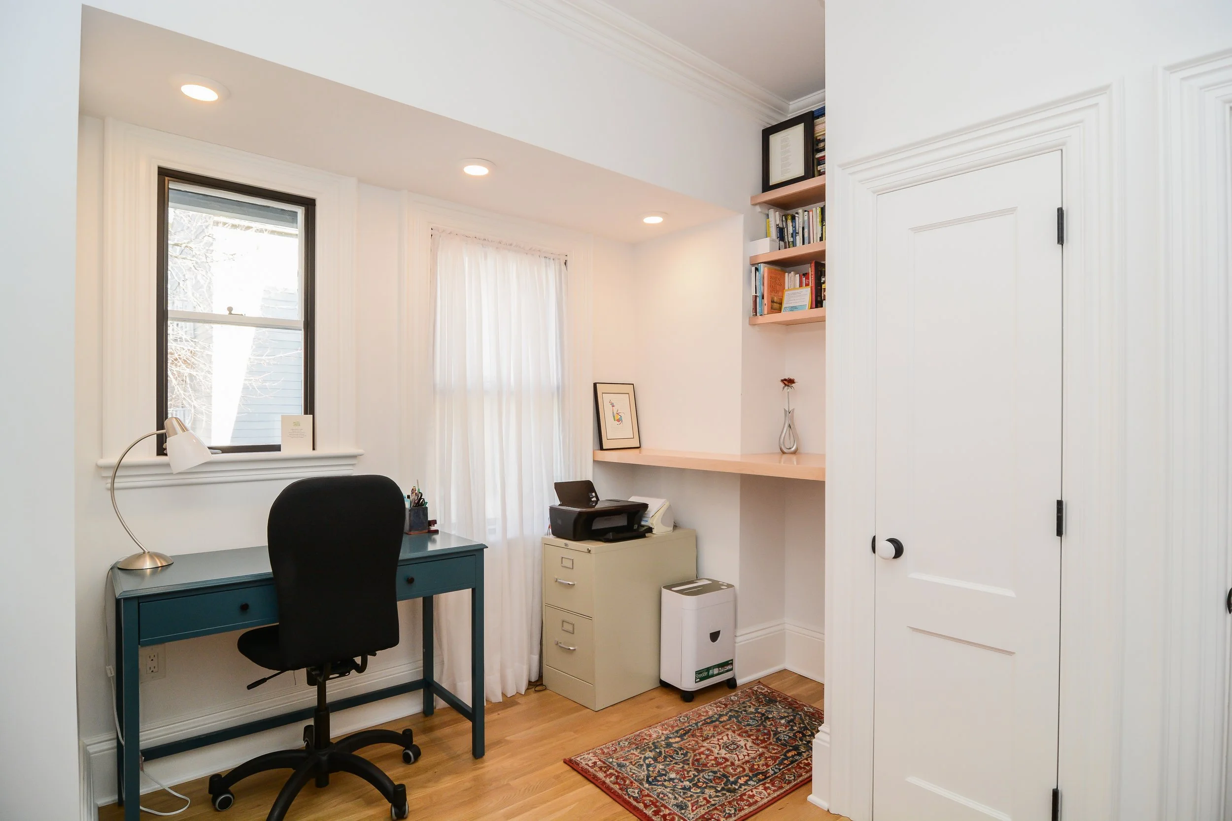 Home office with a blue desk and black chair, window with white curtains, built-in bookshelf with books and a framed picture, beige filing cabinet with a printer, small red patterned rug on hardwood floor.