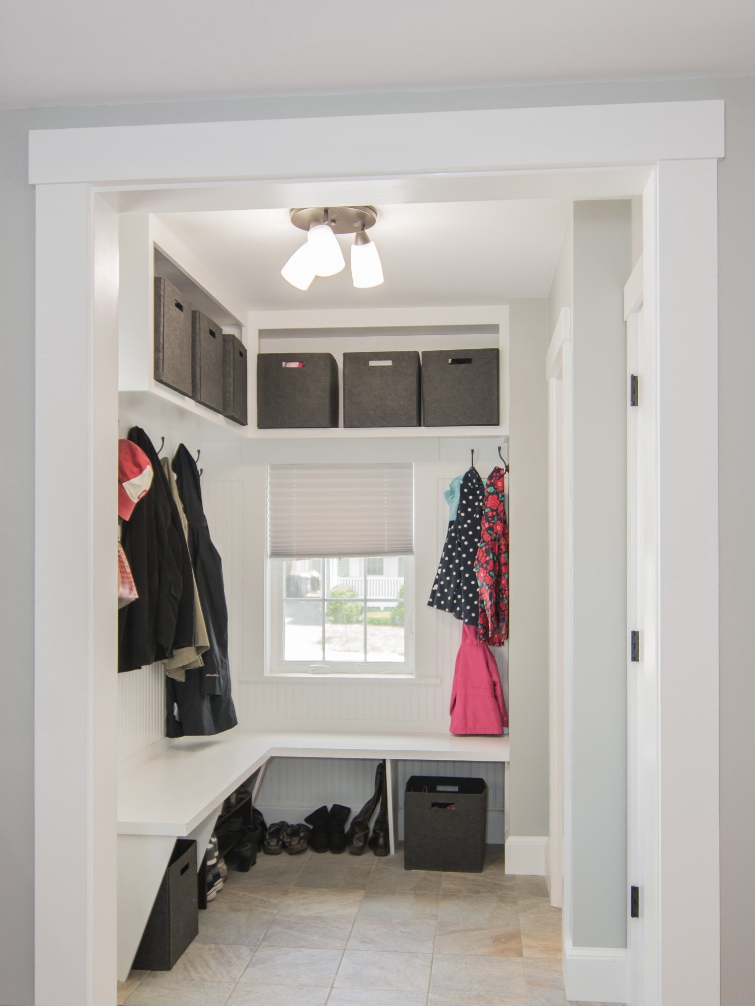A small entryway with a white built-in bench, hooks for hanging jackets, a window with blinds, and wall-mounted shelves with storage boxes at the top. Shoes and a storage bin are on the floor.
