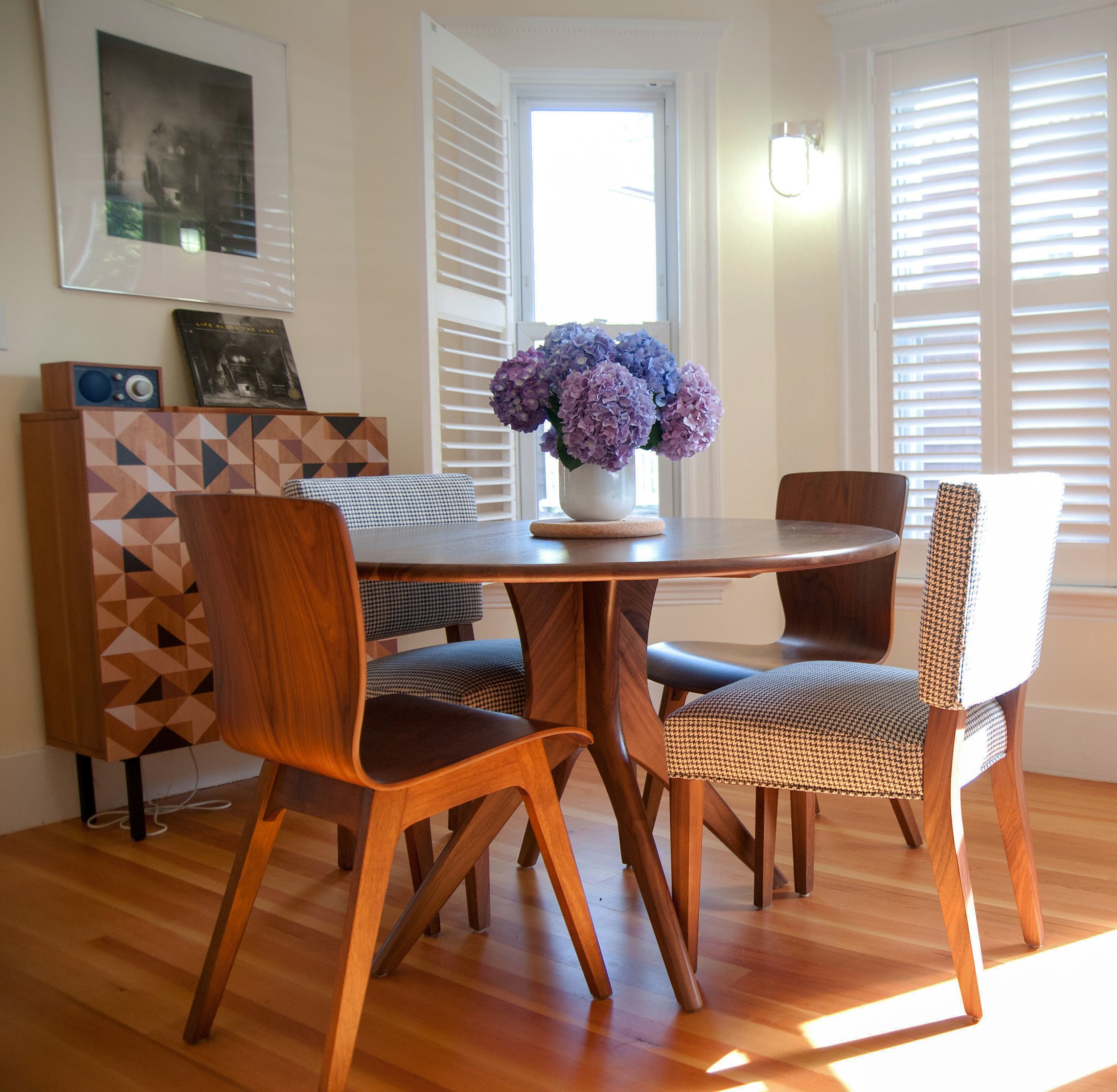 A round wooden dining table with four different style chairs, a white vase with purple hydrangea flowers in the center, and sunlight coming through white window shutters in a bright dining room.