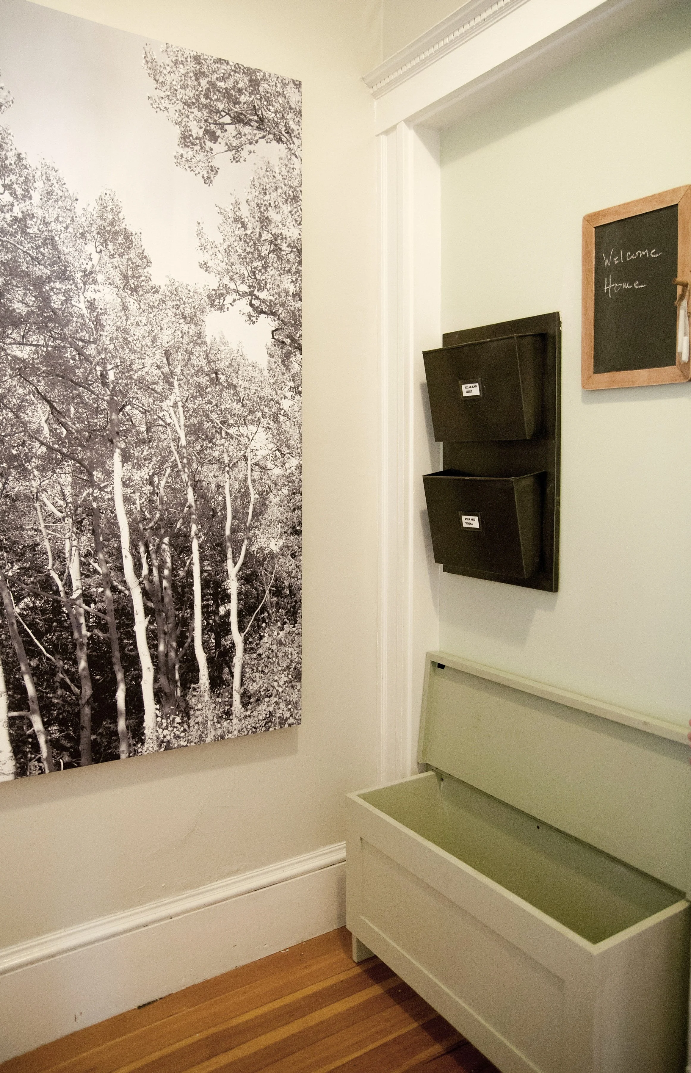 A hallway corner featuring a black and white wall art of trees, two black wall-mounted storage bins, a small blackboard with 'Welcome Home' written on it, and a white bench with an open top, on a hardwood floor.