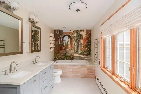 Bright bathroom with dual sinks, large mirrors, a toilet, large window with blinds, and a mural of a Mediterranean street scene on the wall.