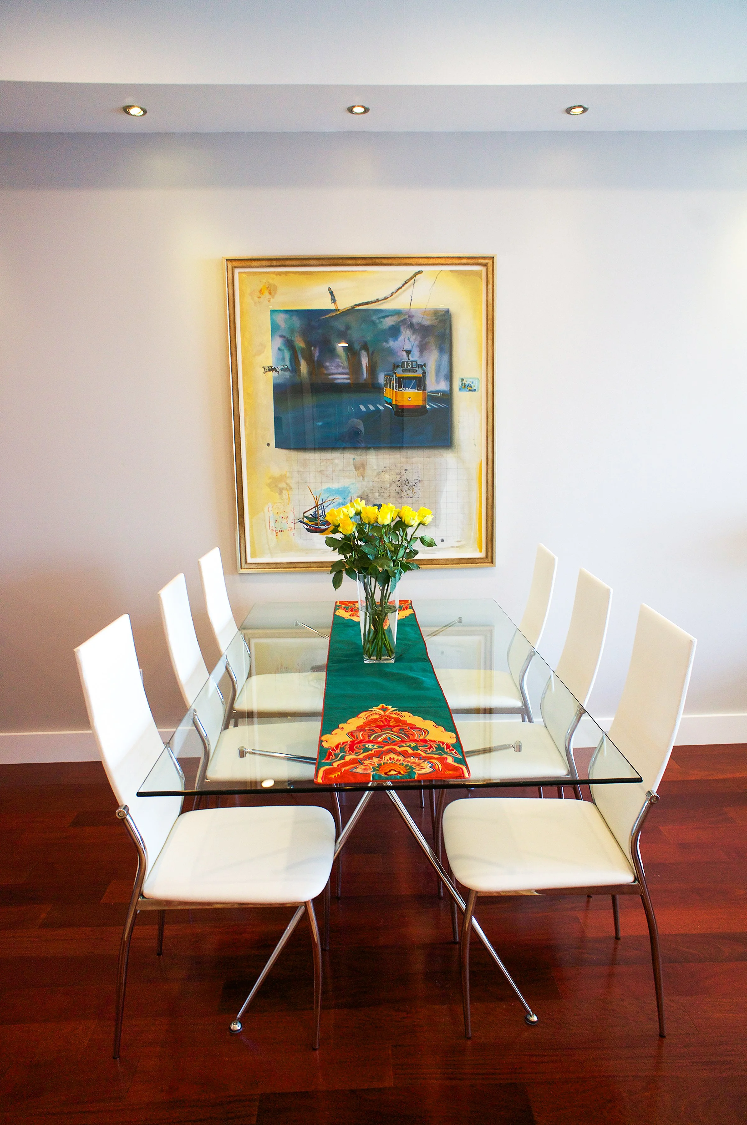A dining room with a glass table, six white chairs, a green and red table runner, a glass vase with yellow roses, and a framed painting of a tram on the wall.