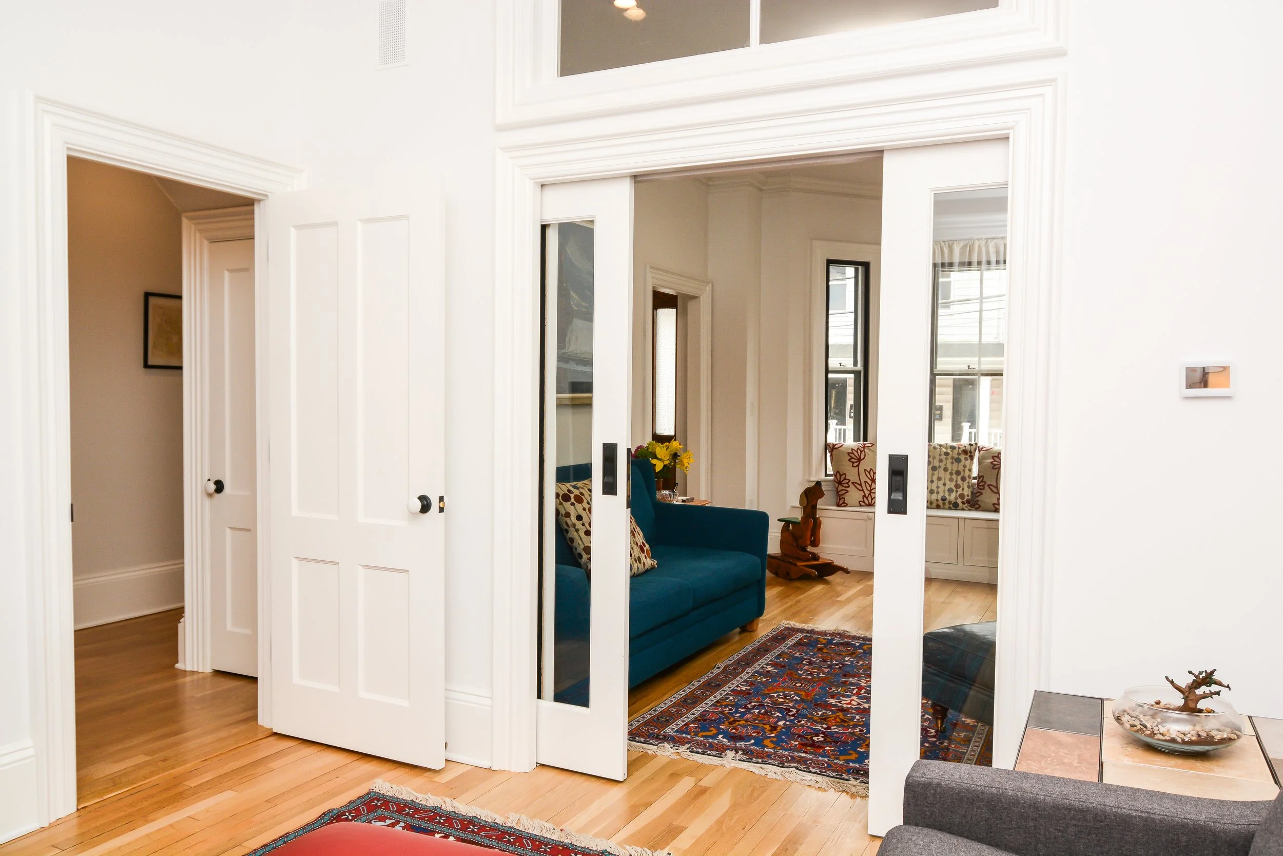 Living room with hardwood floors, white walls, framed picture, and sliding glass doors leading to a room with blue sofa, colorful pillows, window seat, and patterned rugs.