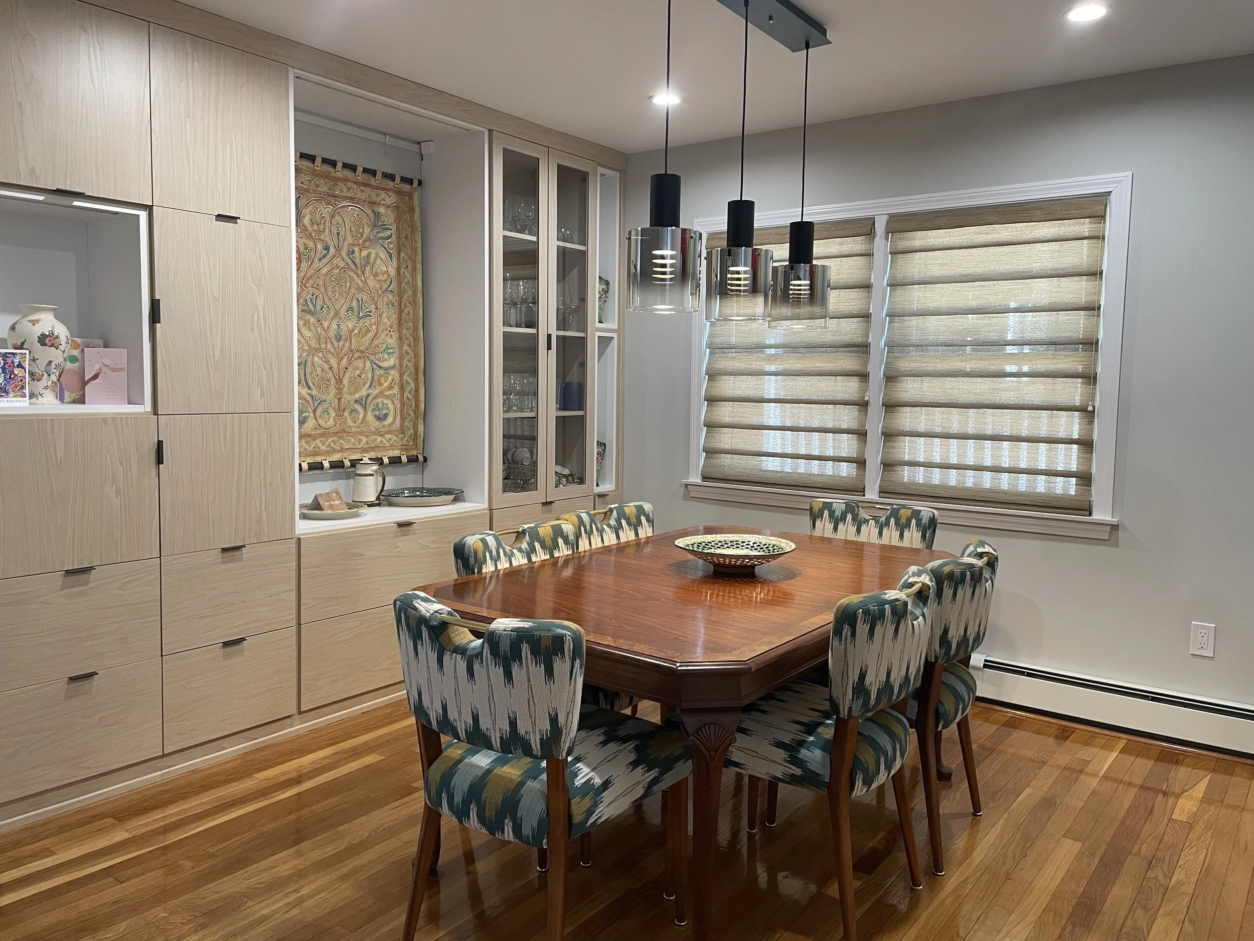 A dining room with a wooden table and six patterned upholstered chairs. There are beige and white window shades and a built-in cabinet with glassware and decorative items. A modern black pendant light fixture hangs over the table.