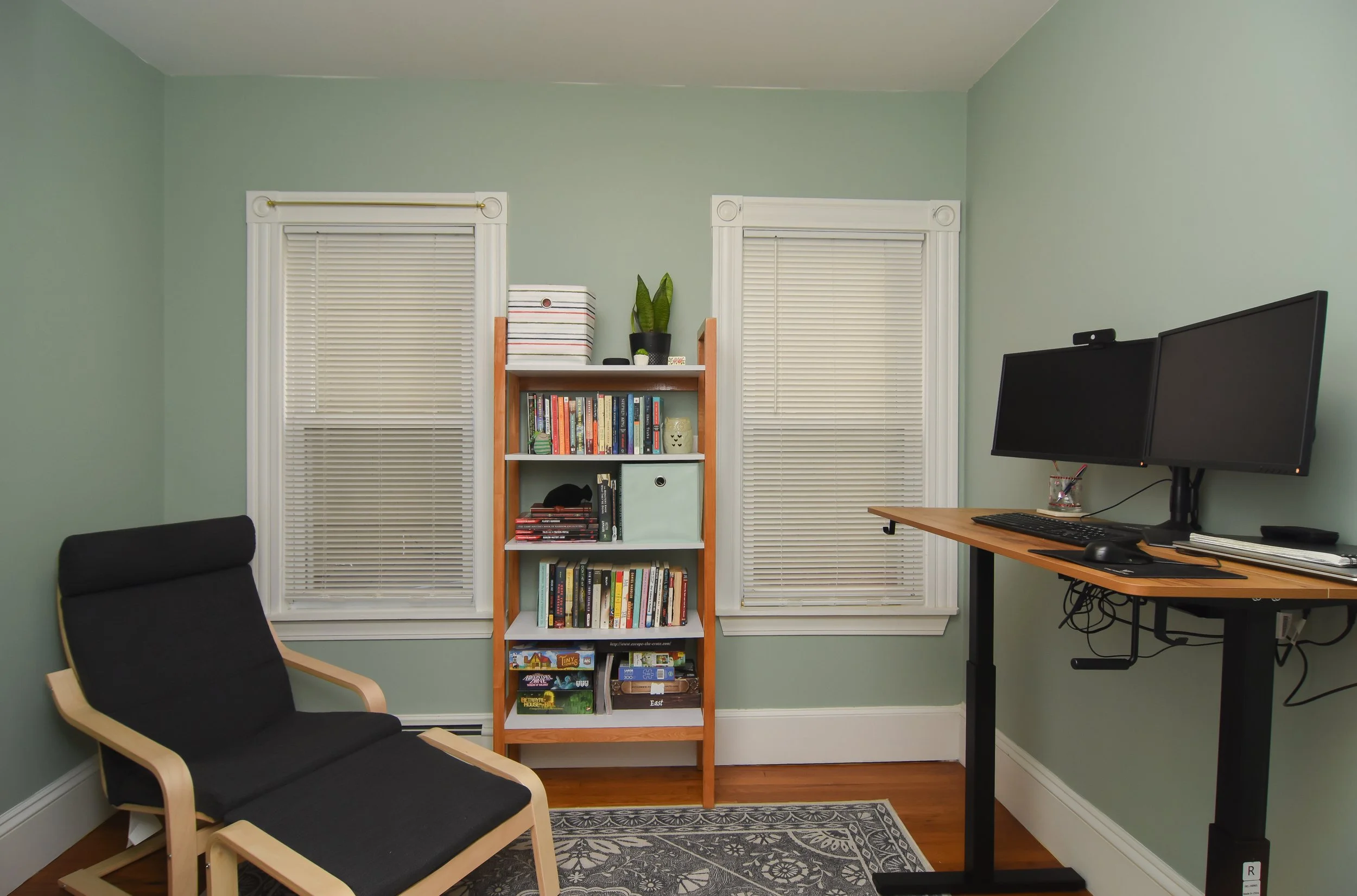 A home office with two large windows with white blinds, a black armchair, a wooden bookshelf filled with books and decorative items, and a standing desk with dual monitors and a keyboard.