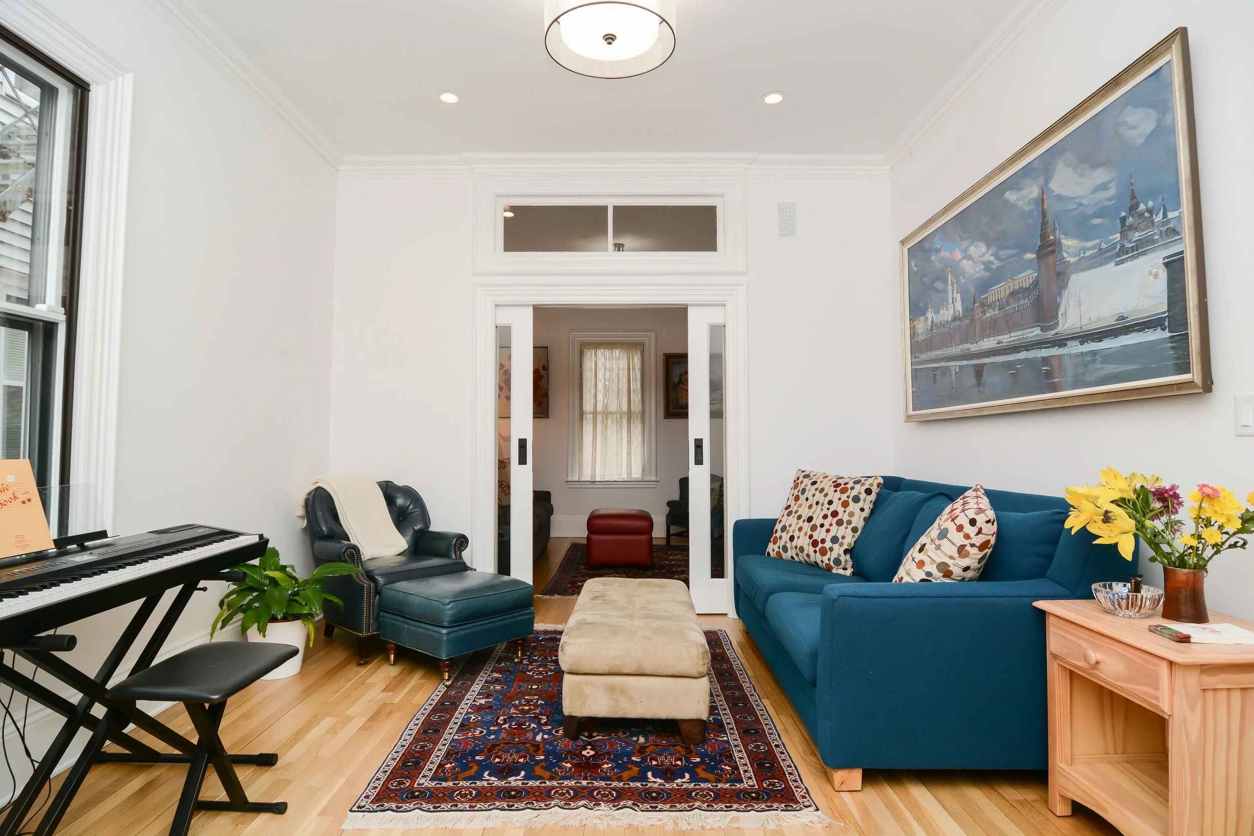 Living room with a blue sofa, patterned pillows, beige ottoman, black leather armchair, green plant, portrait painting of a cityscape, and a wooden side table with fresh flowers.
