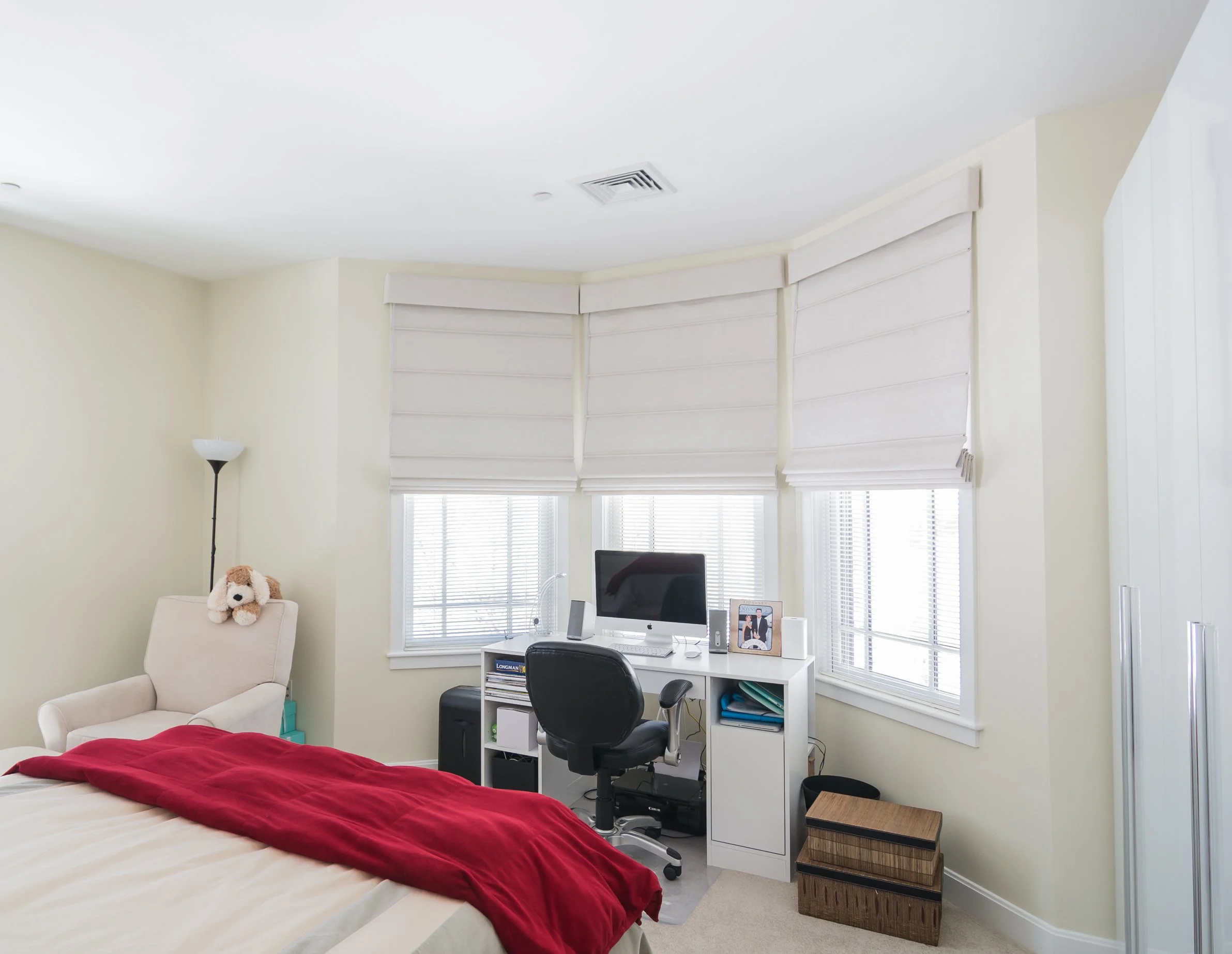 A bright bedroom with a bed, a white desk with a computer, a black swivel chair, a beige armchair with a stuffed animal, a tall lamp, and blinds covering the windows.