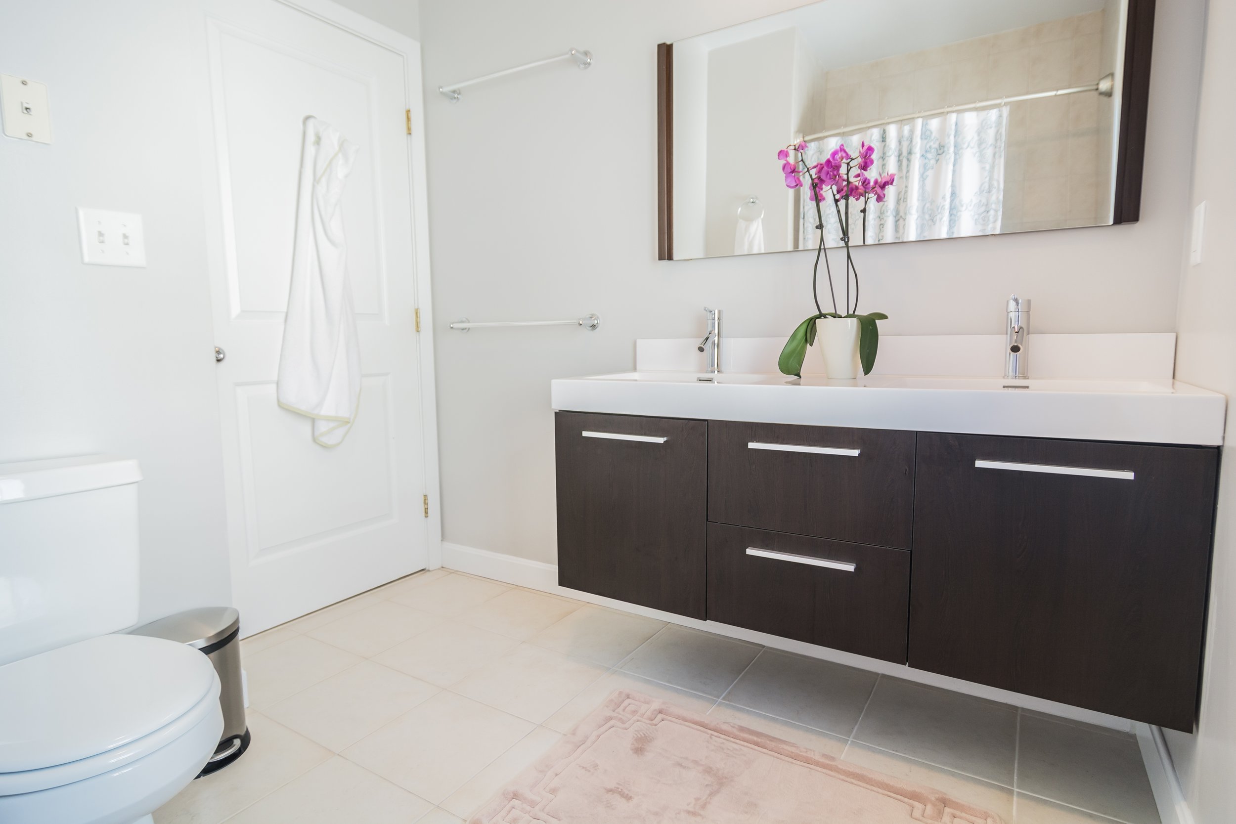 A modern bathroom with a double sink vanity, a large mirror, a potted orchid on the countertop, a white towel hanging on the door, a toilet, a small trash can, and a light-colored tiled floor.