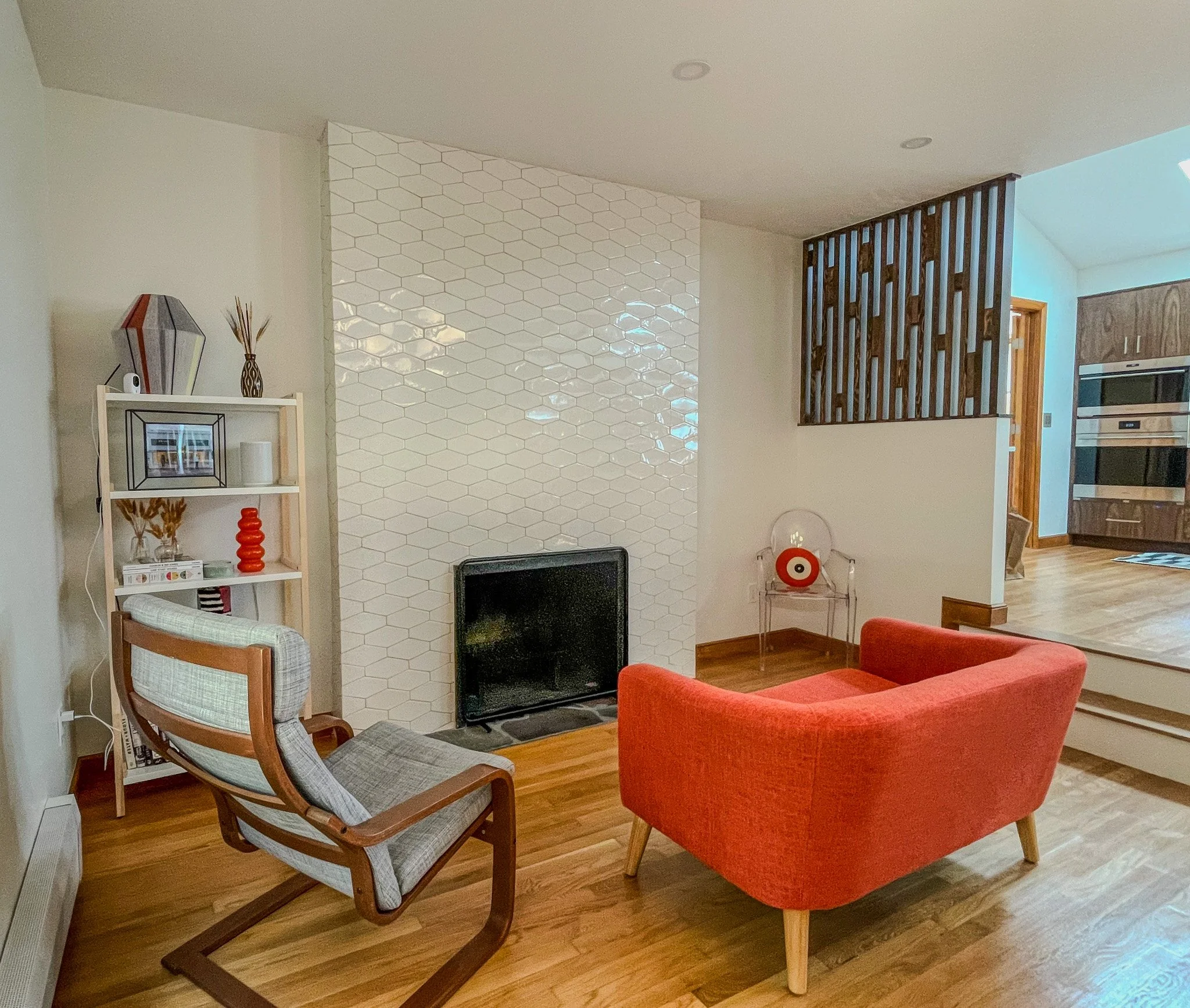 Living room with red armchair, wooden floor, white wall with textured tile wall, bookshelf with decorative items, flat-screen TV, and partial view of kitchen in background
