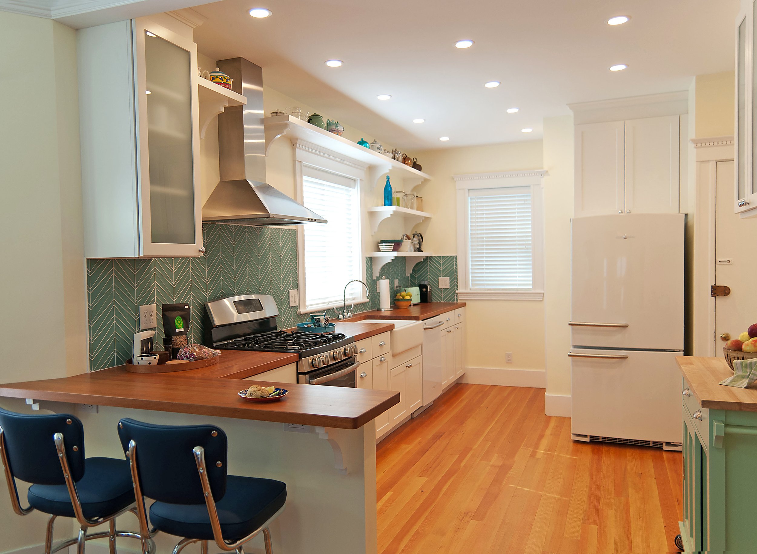 Bright, modern kitchen with white cabinets, wooden countertops, green patterned backsplash, stainless steel stove with oven, white refrigerator, open shelves, and hardwood floors. Two blue chairs are at the kitchen counter.
