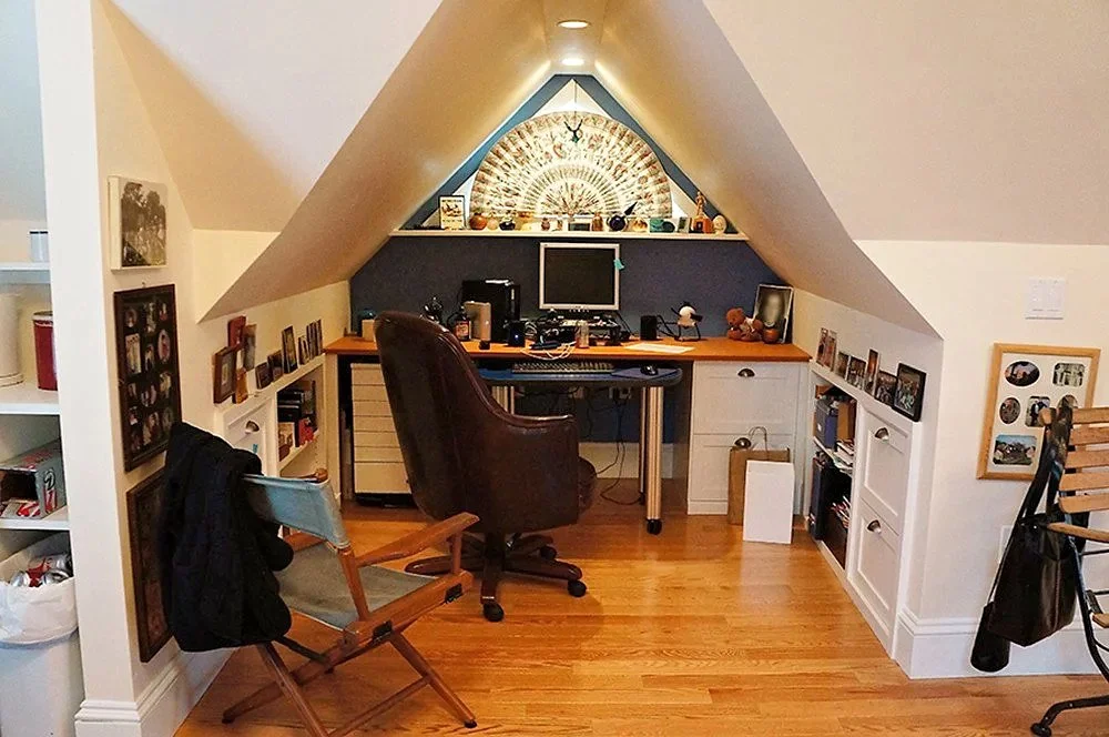Home office space under a sloped ceiling with a wooden desk, black computer monitor, a brown leather chair, and built-in white shelving on both sides decorated with photos and books. A fan-shaped wall decoration with lights is above the desk, and there are various items on the desk and shelves.