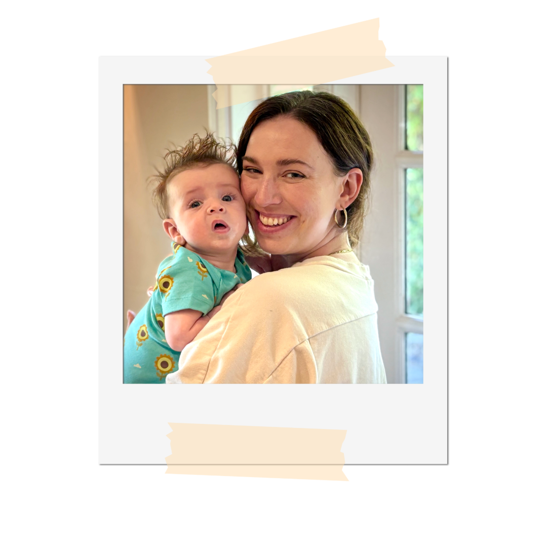 A woman smiling and holding a baby in a room with large windows in the background.