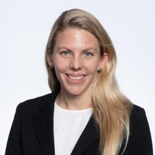 A portrait of a woman with long blonde hair, wearing a black blazer and a white top, smiling against a light gray background.