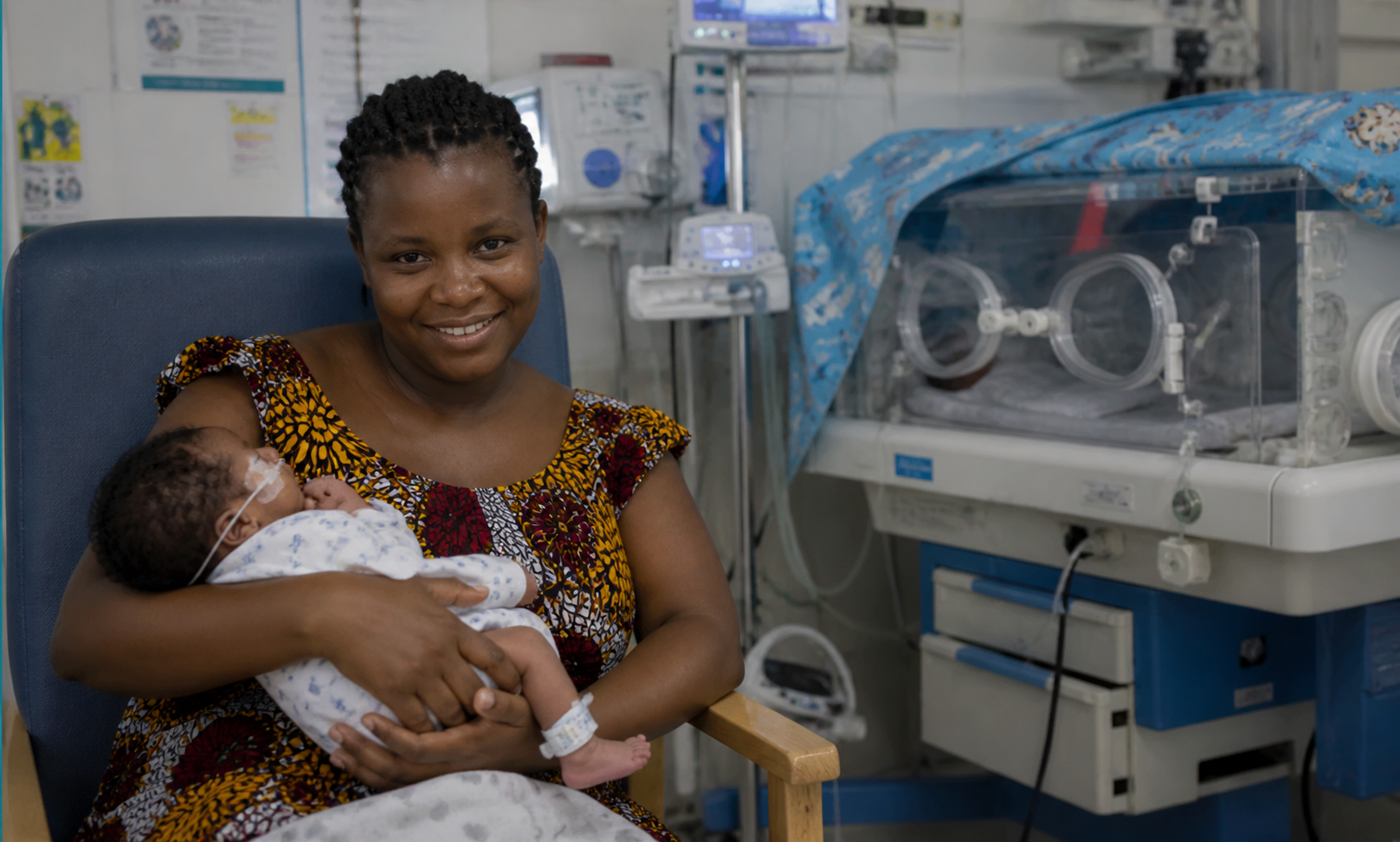 A woman smiling while holding a premature baby in a neonatal intensive care unit, with medical equipment and incubators in the background. CPAM support.