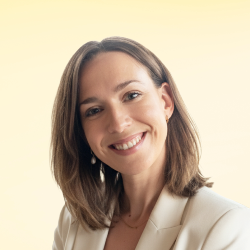 A smiling woman with shoulder-length brown hair, wearing a white blazer and pearl earrings, against a light yellow background.