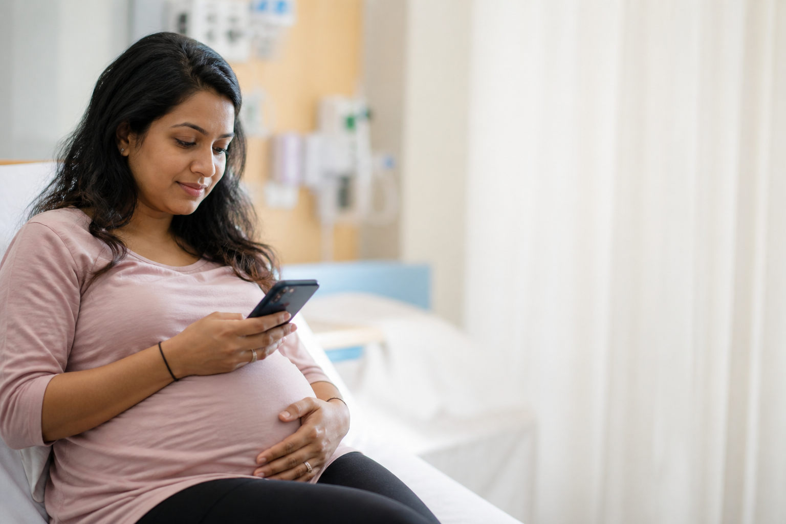 Pregnant woman sitting in a hospital room, looking at her phone and cradling her baby bump. CPAM baby support.