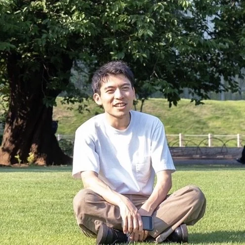 芝生の上に座る若い男性、背景は木と緑の景色、晴れた日