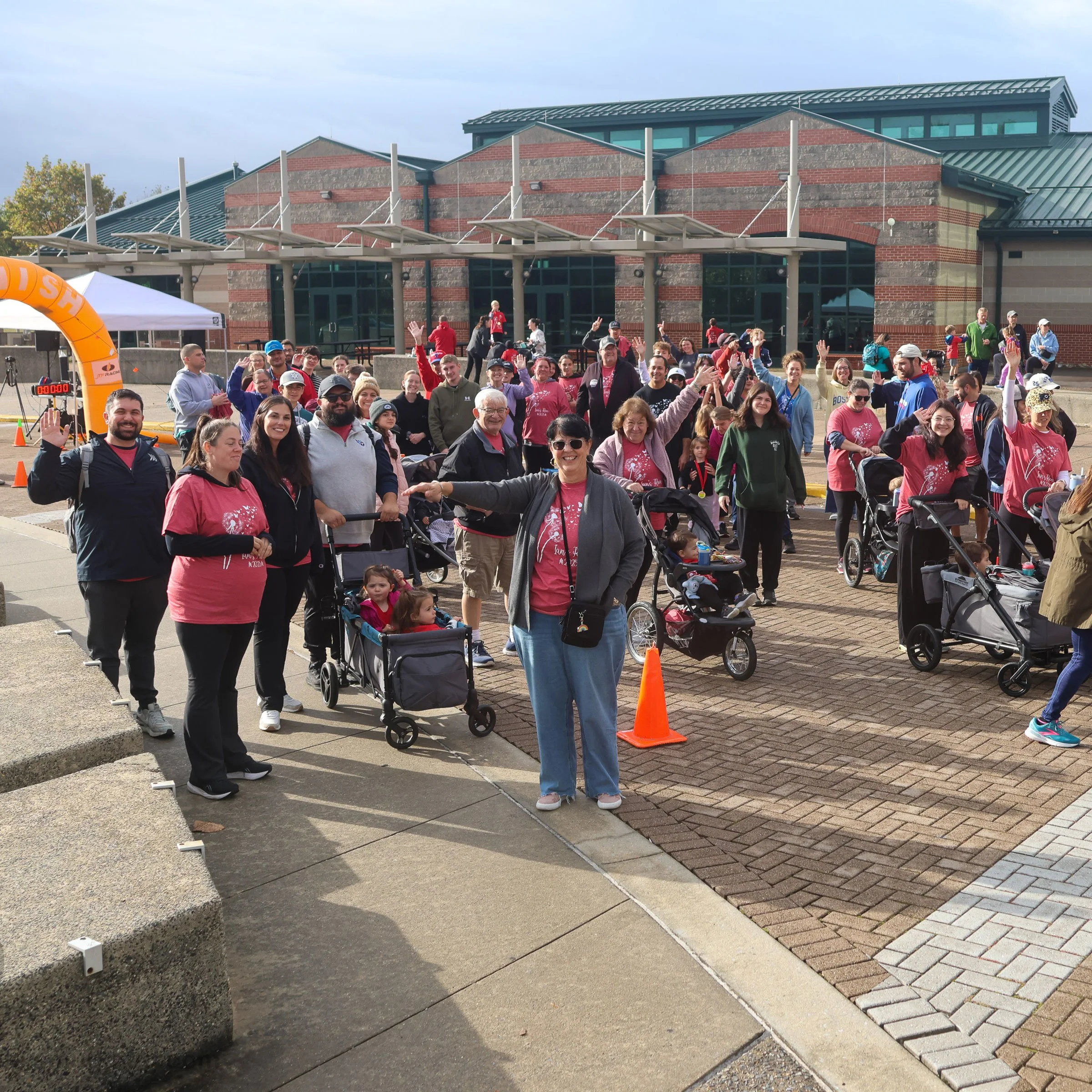 A large group of people gathered outdoors for a walk or event, many with strollers, wearing pink shirts, with a building in the background and an orange inflatable arch on the left.