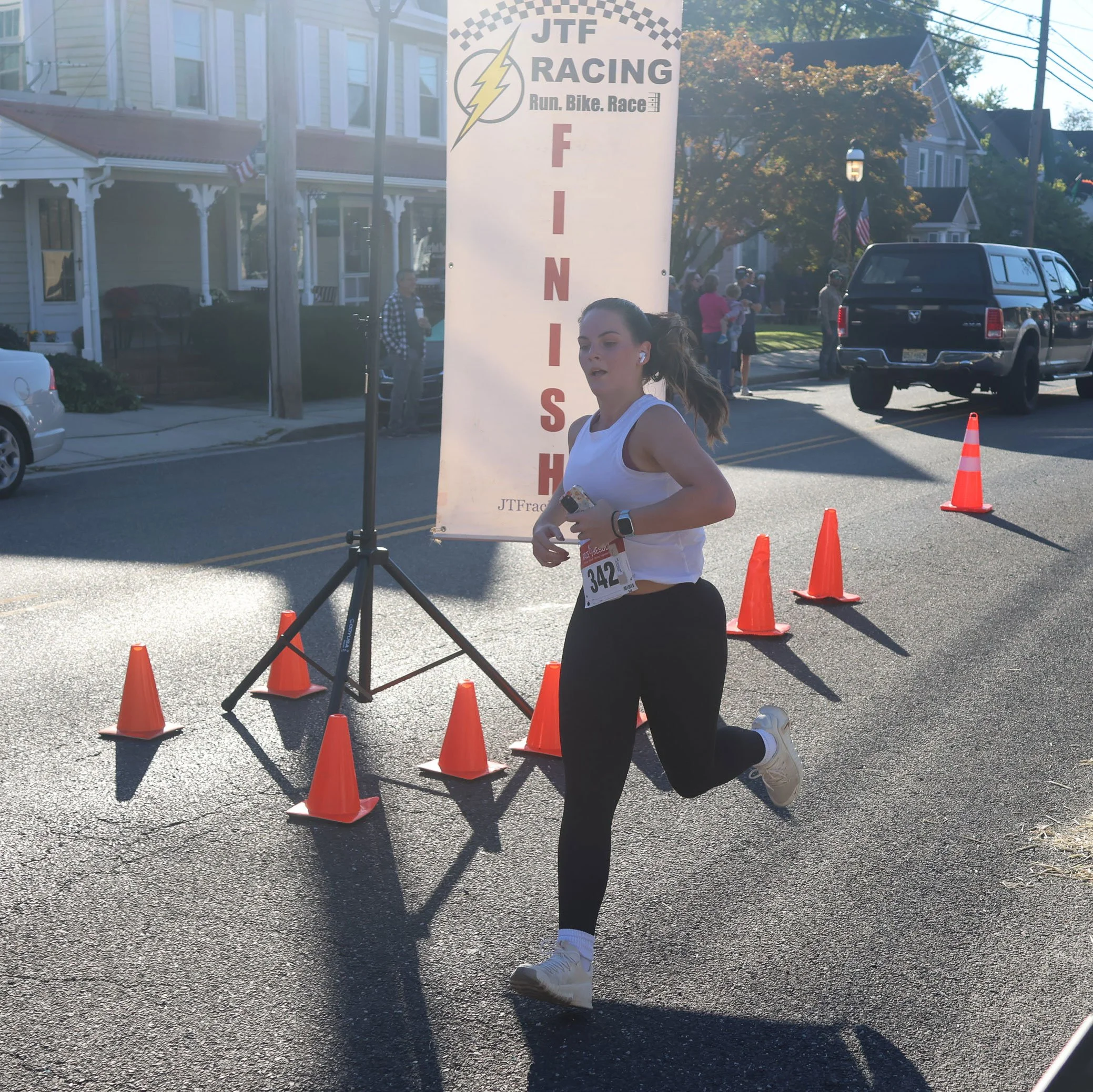 A young woman with a running bib and a smartwatch runs past a finish line at a race, with a banner that reads 'JTF Racing' and 'Finish' in the background, on a sunny street lined with houses and cars.
