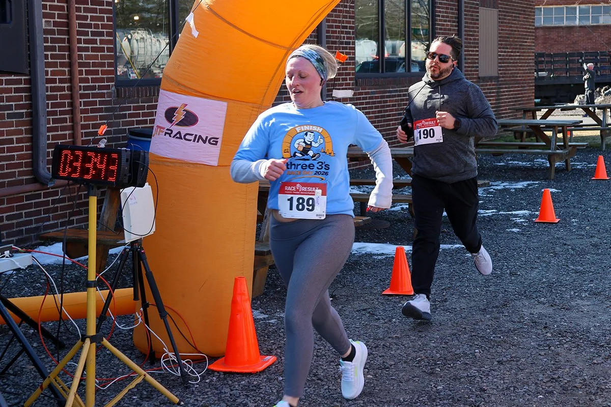 Female runner finishing the race wearing the Three 3's Race Day t-shirt and a race bib.