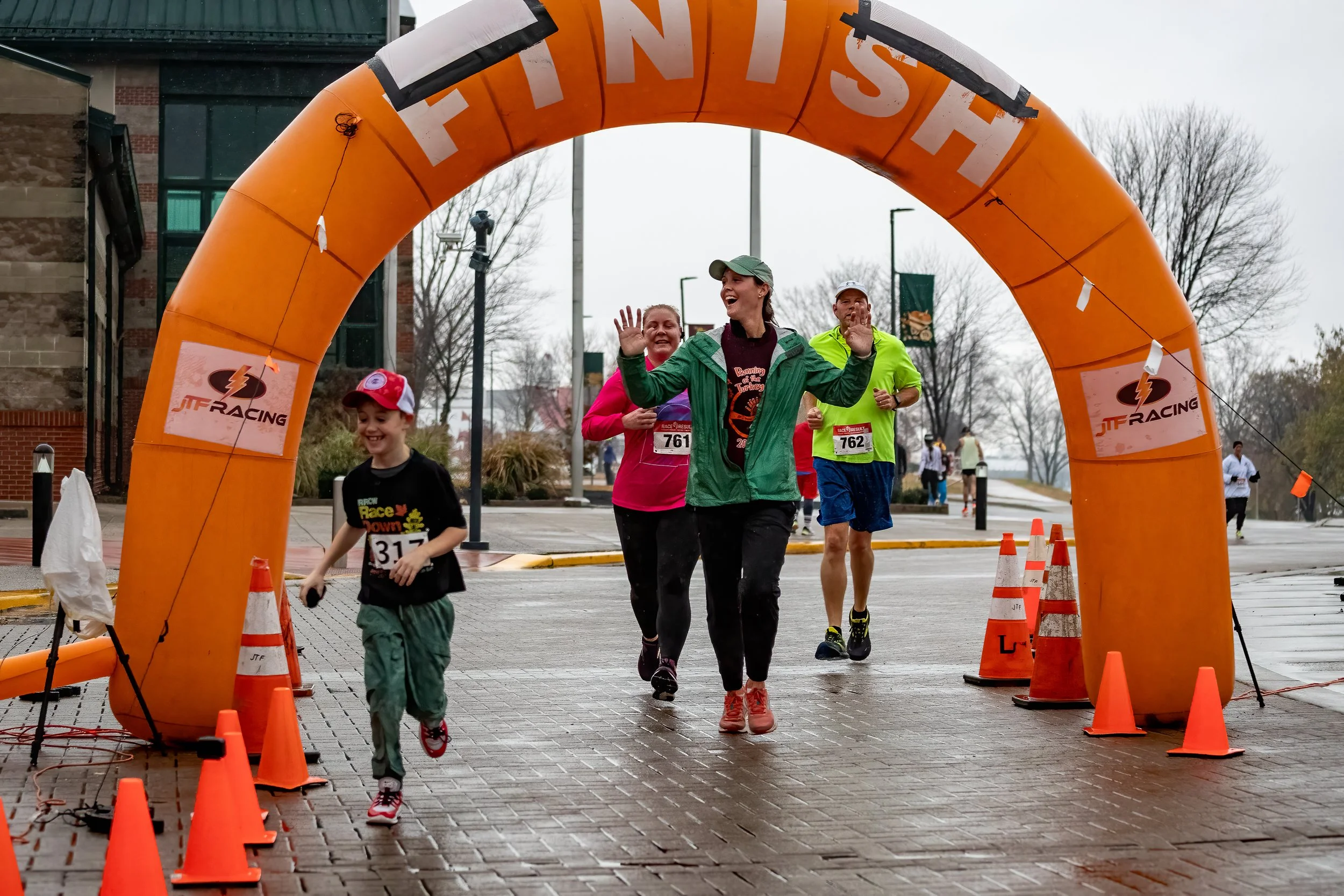 People crossing a finish line at a race under a large orange arch with the word 'FINISH' on it, on a cloudy day, with orange cones on the ground.