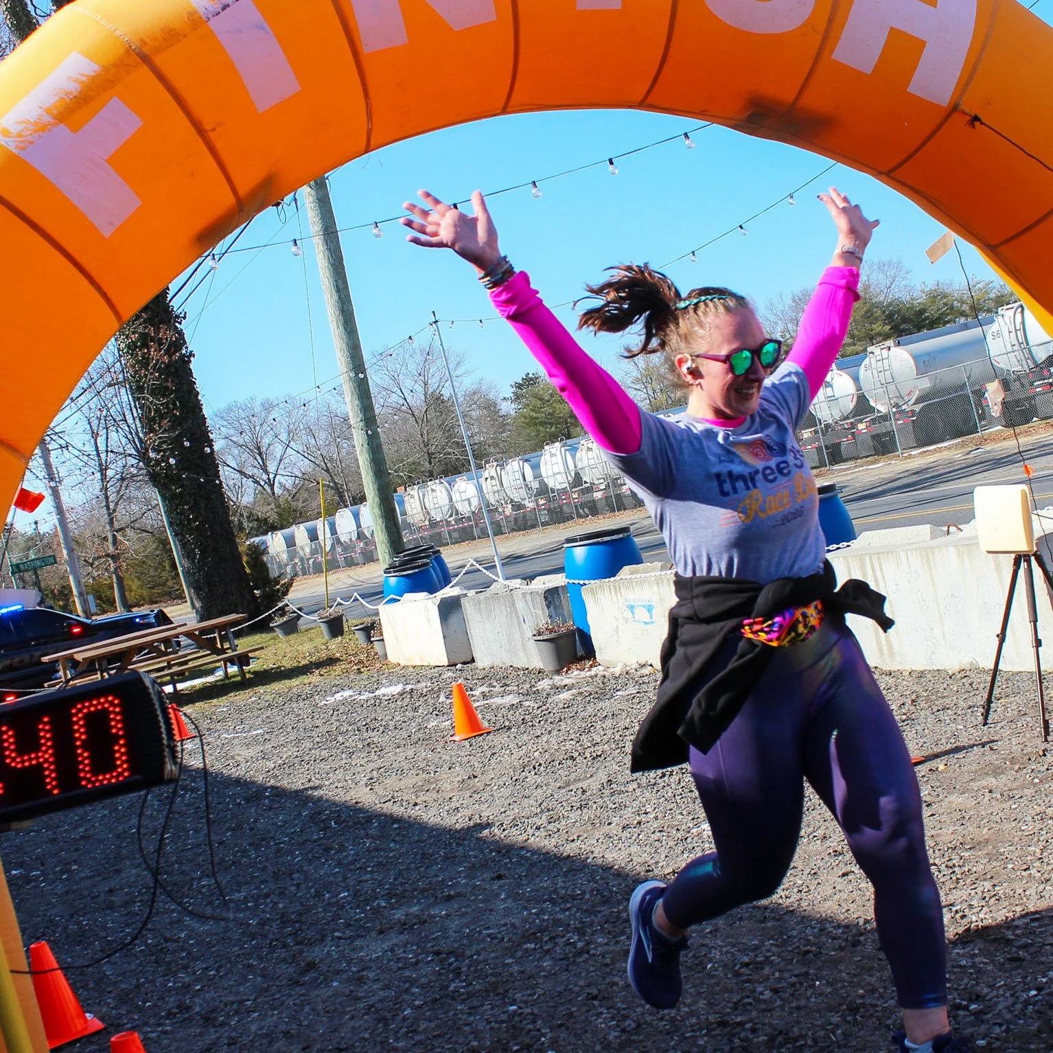 A woman wearing sunglasses and a race shirt crosses the finish line of a race with arms raised in celebration.