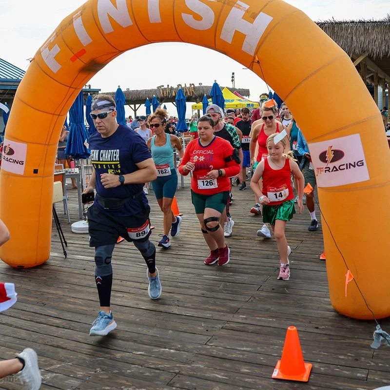 People participating in a running race, starting under an orange inflatable arch that says 'FINISH' in large white letters, on a wooden boardwalk at an outdoor event.