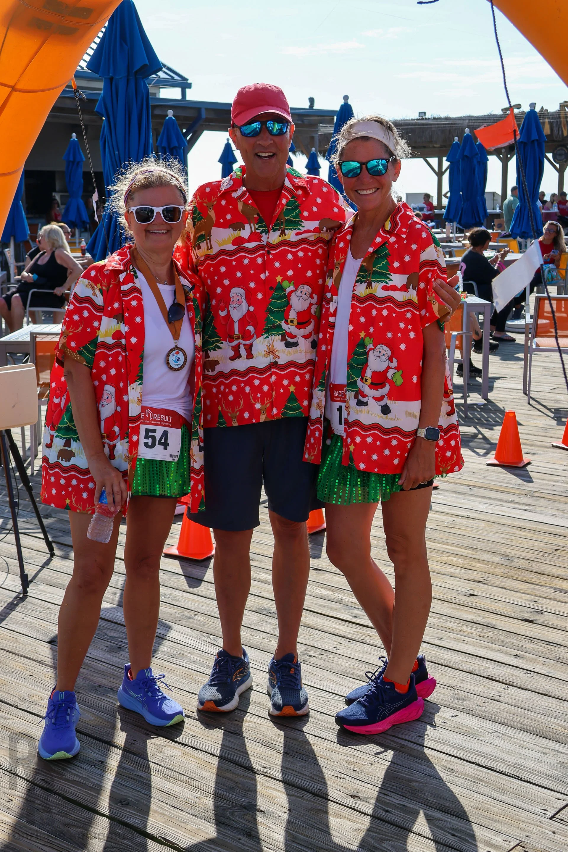Three smiling people in festive Christmas shirts standing together outdoors on a wooden deck with blue umbrellas and other people in the background.